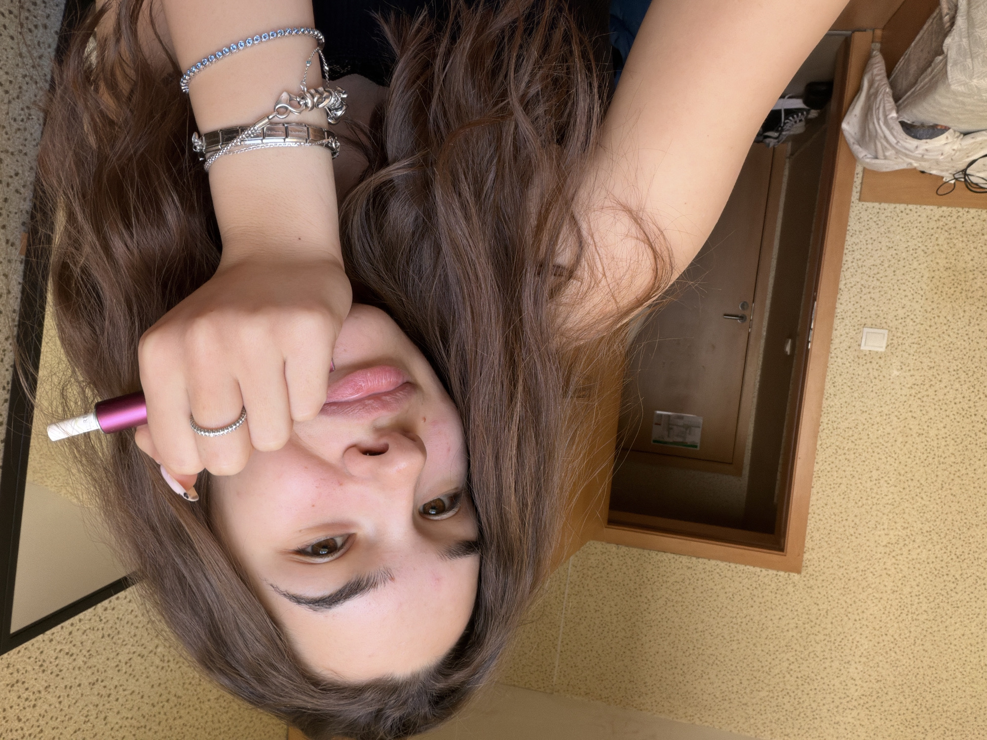 The image displays a close-up, slightly tilted portrait of a young person, likely female, with long, wavy brown hair framing her face. She has fair skin, dark brown eyes, and appears to be looking directly at the camera with a neutral expression, her lips slightly parted.
Her left arm is visible, adorned with several silver bracelets: one featuring small blue gemstones, another with intricate silver charms (including one resembling a stylized animal or knot), and a wider, flat-linked bracelet. On her right ring finger, she wears a silver ring embellished with small stones. She is holding a slim, cylindrical object, purple with a white tip, near her mouth with her right hand. Faint text, "PURI", is visible on the white tip of the object, which could be a vape pen or a cosmetic tool.
The background suggests an indoor setting, possibly a room in a residence in Sofia, Bulgaria. A wooden door frame and a dark interior are visible to the right and top. The wall features a light-colored, speckled wallpaper or textured paint, and a white light switch is mounted on it. Some household items, including what appears to be folded clothes and wires, are visible on a surface near the door frame. The lighting is soft and even, typical of an indoor environment, making the time of day indistinguishable.