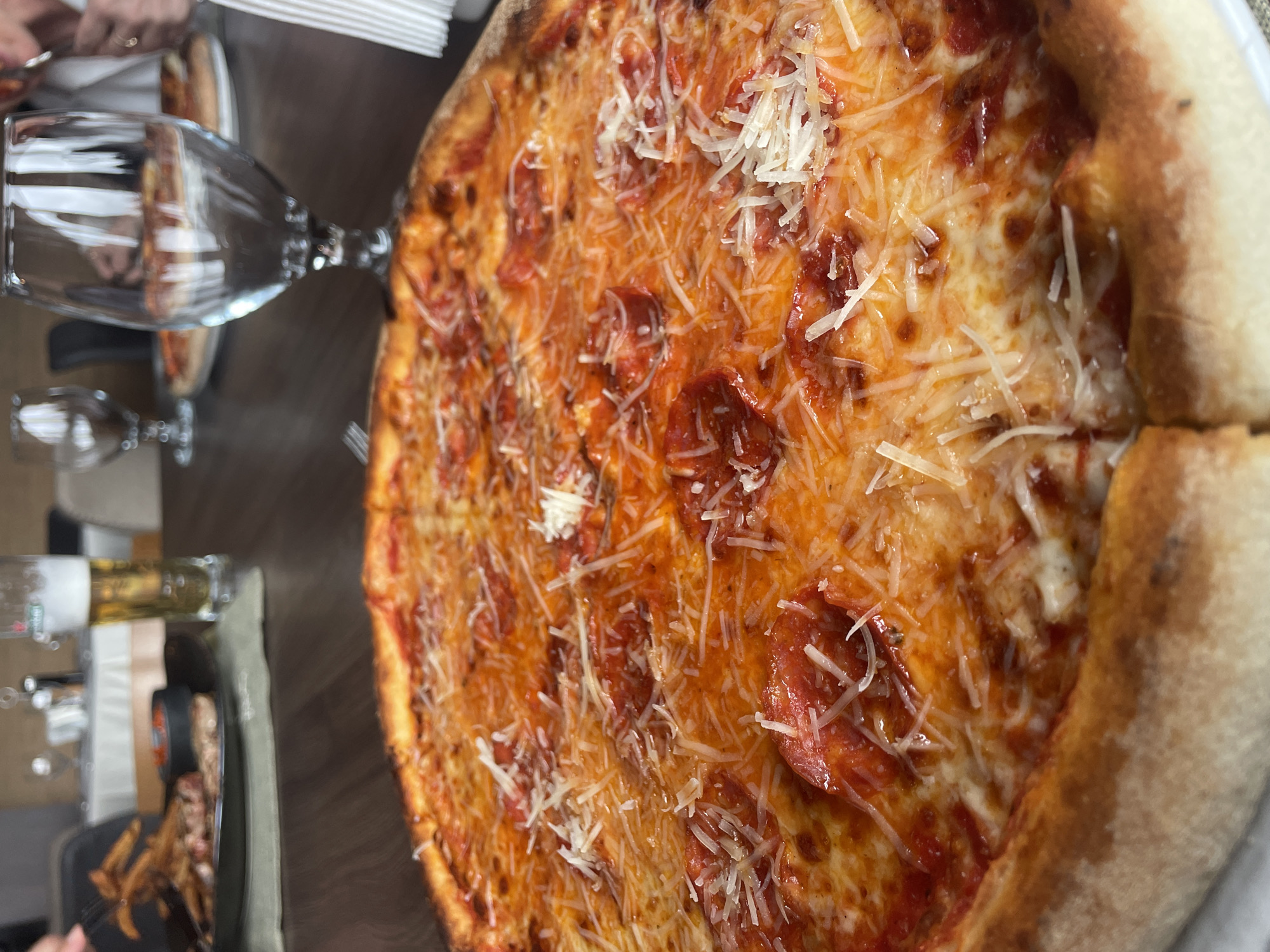 The image shows a pepperoni pizza on a table. The pizza is topped with red sauce, pepperoni slices, and shredded cheese. A glass of wine is also on the table. There are french fries in a dish on the left. The location in the city of Pleven, Bulgaria is not determinable from the provided information.