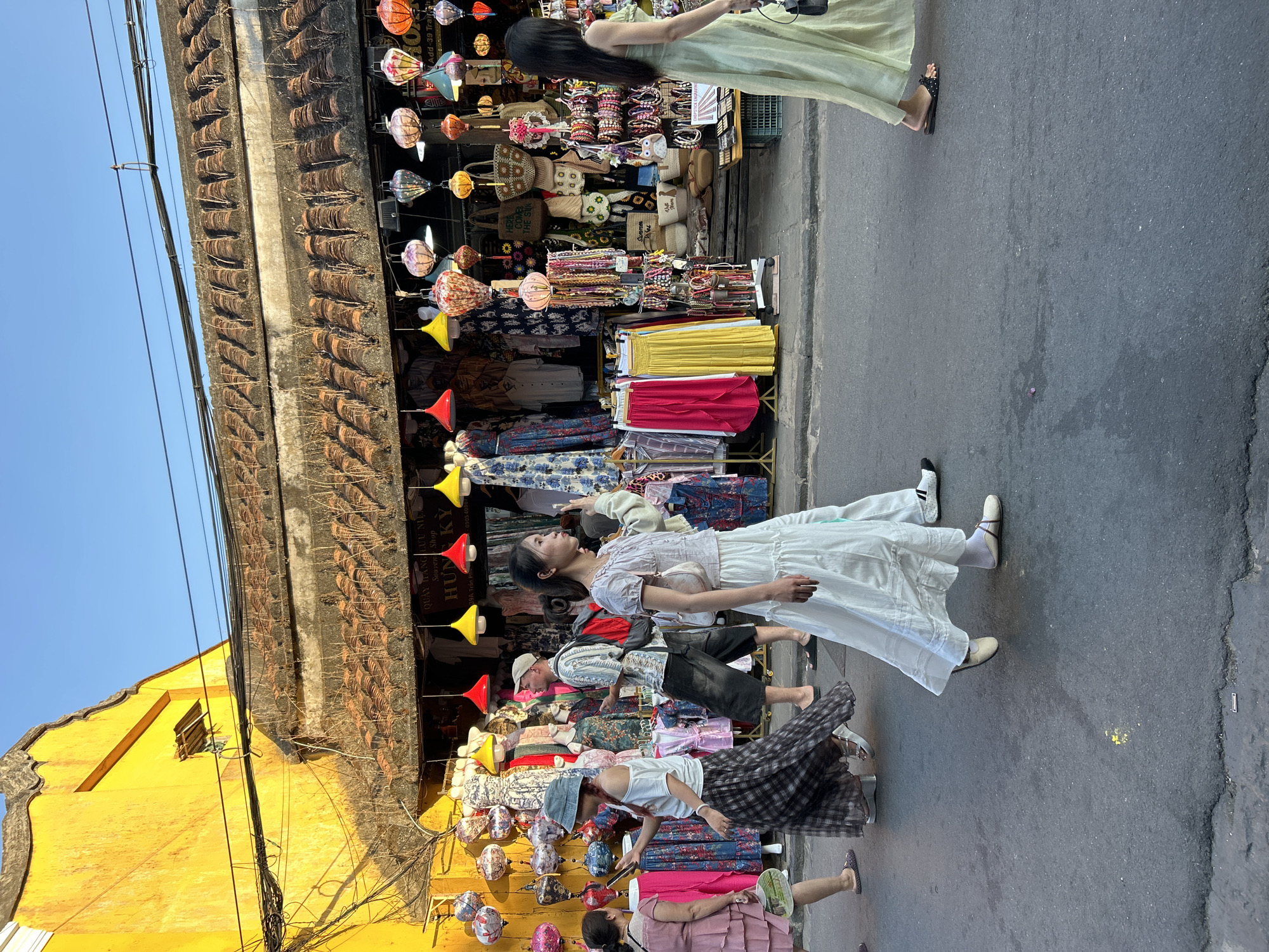 In a street scene in Hoi An Ward, Vietnam, several people walk past a brightly lit shop displaying a vibrant array of goods, including clothing and lanterns. The shop's facade is painted a striking yellow, with a tiled roof and an overhang draped with colorful lanterns of various shapes and sizes. In the foreground, a woman in a flowing light green dress walks to the right, her side profile visible as she holds a dark handbag. To her left, a woman in a white top and long white skirt walks towards the right, her hand raised slightly. Further left, another woman in a blue hat and plaid skirt walks to the right, looking at her phone. A man in a light-colored patterned shirt and dark shorts is also visible, walking to the right with a red backpack. A woman in a pink dress is seen on the far left, walking to the right with a fan. The street is paved with asphalt, and the overall atmosphere suggests a bustling market or shopping area in the city.