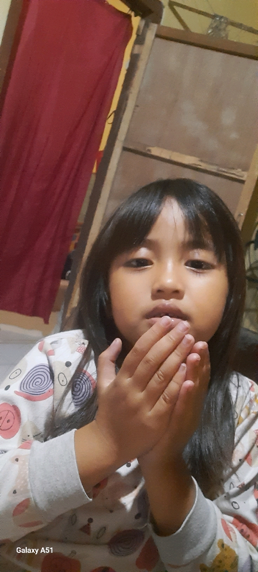 A young Asian girl, estimated to be between 4-6 years old, is in a medium shot, facing the camera and holding her hands together near her mouth. She has long, dark hair that falls to her shoulders, bangs framing her forehead, and dark eyes. She is wearing a light-colored pajama top with a pattern of cartoon-like animals and swirls. Her hands are clasped in front of her mouth, with her fingers slightly splayed. The background is dimly lit and appears to be an indoor setting. To the left of the girl, a large, dark red curtain hangs, partially obscuring a wooden structure. To the right of the girl, the wooden structure continues, with a lighter, possibly plywood surface visible. The overall impression is of a casual, home environment. The bottom left corner of the image displays the text "Galaxy A51," indicating the device used to capture the photograph, and the location "Ploso, Indonesia."