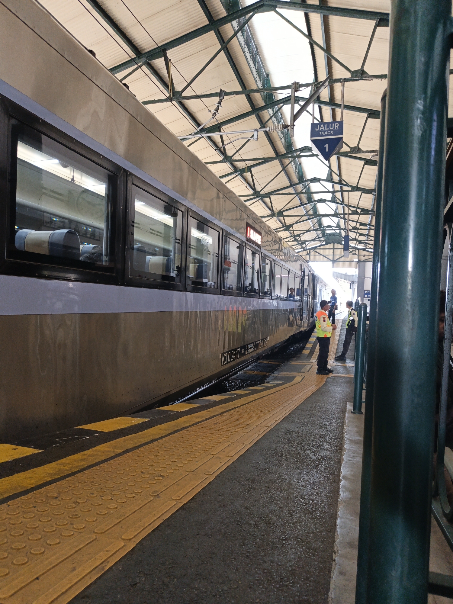 The image depicts a train at a station, identified as Klaten Station in Klaten, Indonesia. A silver and beige train is parked alongside a platform. The platform has a yellow tactile paving surface and yellow safety lines. Two individuals wearing high-visibility vests and dark trousers are standing on the platform, appearing to be station personnel. One individual is facing the train, while the other is looking towards the first individual. A blue sign with white text reading "JALUR TRACK 1" is visible hanging from the station's roof structure, indicating Track 1. The station has a covered roof supported by metal beams. The environment appears to be outdoors, with natural light filtering through the roof.
