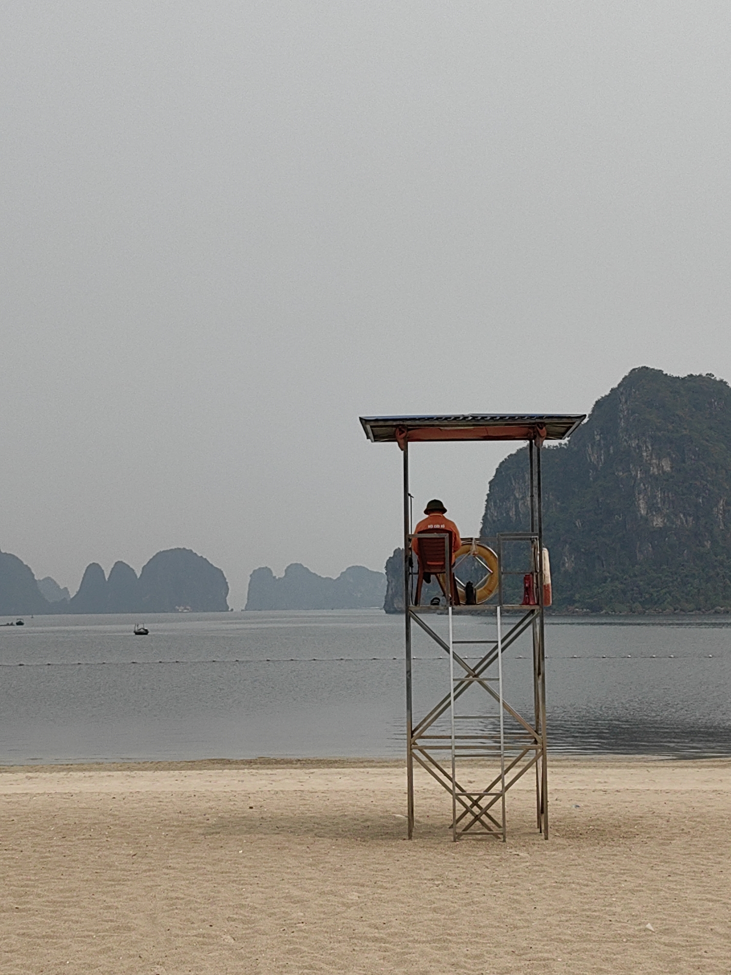 The image captures a quiet coastal scene, likely in Ha Long Ward, Vietnam, characterized by the distinctive karst limestone islands rising from the sea. In the foreground, a sandy beach stretches out, showing faint ripple marks.
The central subject is a lifeguard seated in a tall, metal observation stand, viewed from behind. The lifeguard wears an orange vest with Chinese characters "救护员" (jiùhùyuán), meaning "lifeguard" or "rescuer," printed on the back, and a dark hat. They are positioned on a red chair, looking out towards the water and the distant islands. A yellow lifebuoy is visible, hanging within the structure of the stand.
Beyond the beach, the calm, gray waters of the bay are dotted with a few small boats and marked by a line of buoys. In the mid-ground and background, numerous dark, verdant karst islands emerge from the water under an overcast sky. The weather appears hazy or cloudy, contributing to a muted color palette and soft, diffused light across the entire scene. The time of day appears to be daytime, under subdued light conditions. The overall atmosphere is one of calm vigilance.