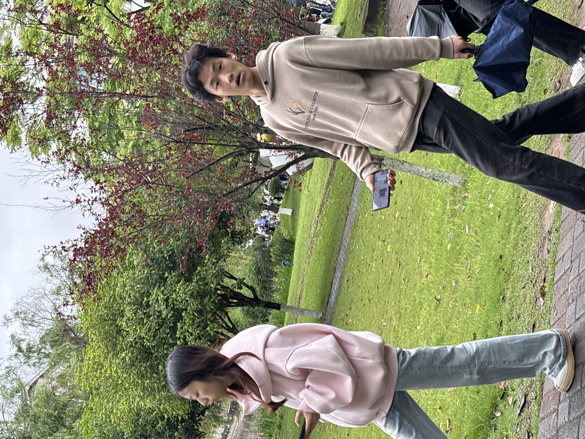 Two people are walking on a brick path in a park. The man, wearing a tan hoodie, black pants, and holding an umbrella and a phone, is walking alongside the woman. She is wearing a pink hoodie and light blue jeans, and appears to be looking down at something. The location is a park in the city of Jiangjin, China. Trees and green grass can be seen in the background.