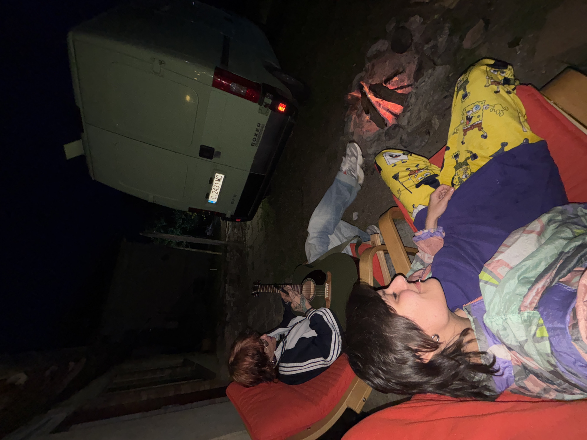 The image depicts a nighttime scene in Karlukovo, Bulgaria. A light-colored Peugeot Boxer van is parked next to a campfire. Two people are seated in red lounge chairs. One person is wearing SpongeBob SquarePants pajamas, covered with a purple blanket. Another person is playing a guitar. A pair of jeans and a white sneaker are lying on the ground. The background includes a dark structure, possibly a building.