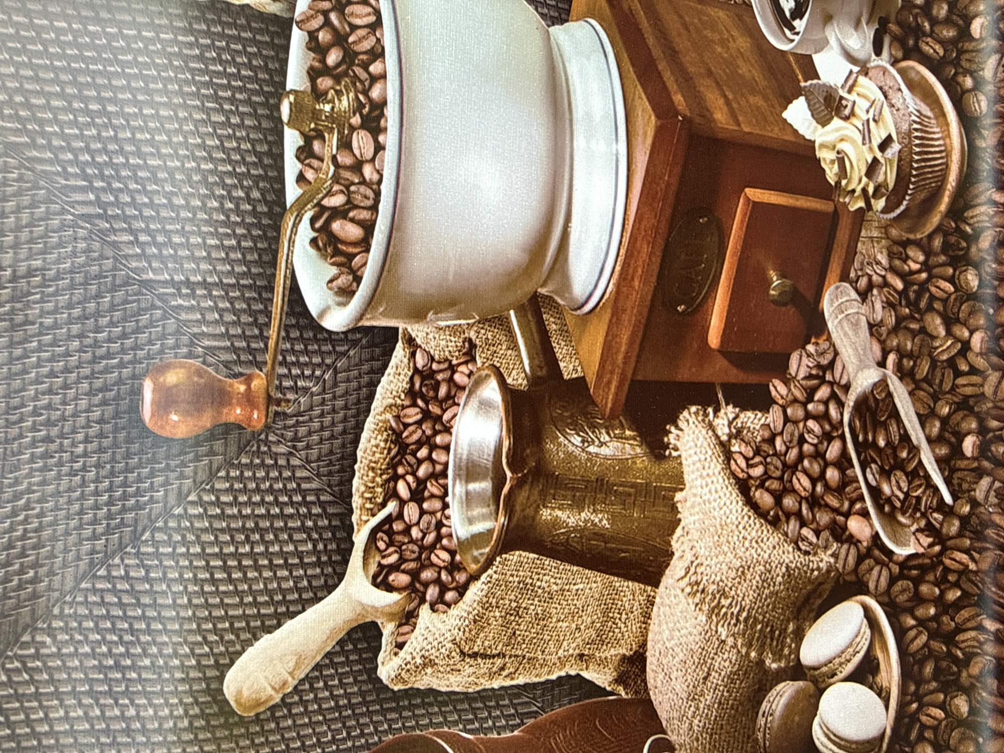 The image depicts a still life arrangement centered around coffee-related items. In the background, a textured, woven surface creates a backdrop. Prominently featured are a burlap sack overflowing with roasted coffee beans, a wooden scoop resting within the sack, and a copper cezve (Turkish coffee pot) placed in front of it. To the right, a white bowl filled with coffee beans sits atop a wooden coffee grinder, with a golden crank extending from it. Further to the right, a cupcake decorated with frosting and chocolate shavings is visible, next to a cup of coffee. Scattered throughout the scene are more coffee beans, along with two macarons on a small plate. The overall impression is one of a cozy coffee-themed composition, with no discernible connection to the city of Bilenke, Ukraine.