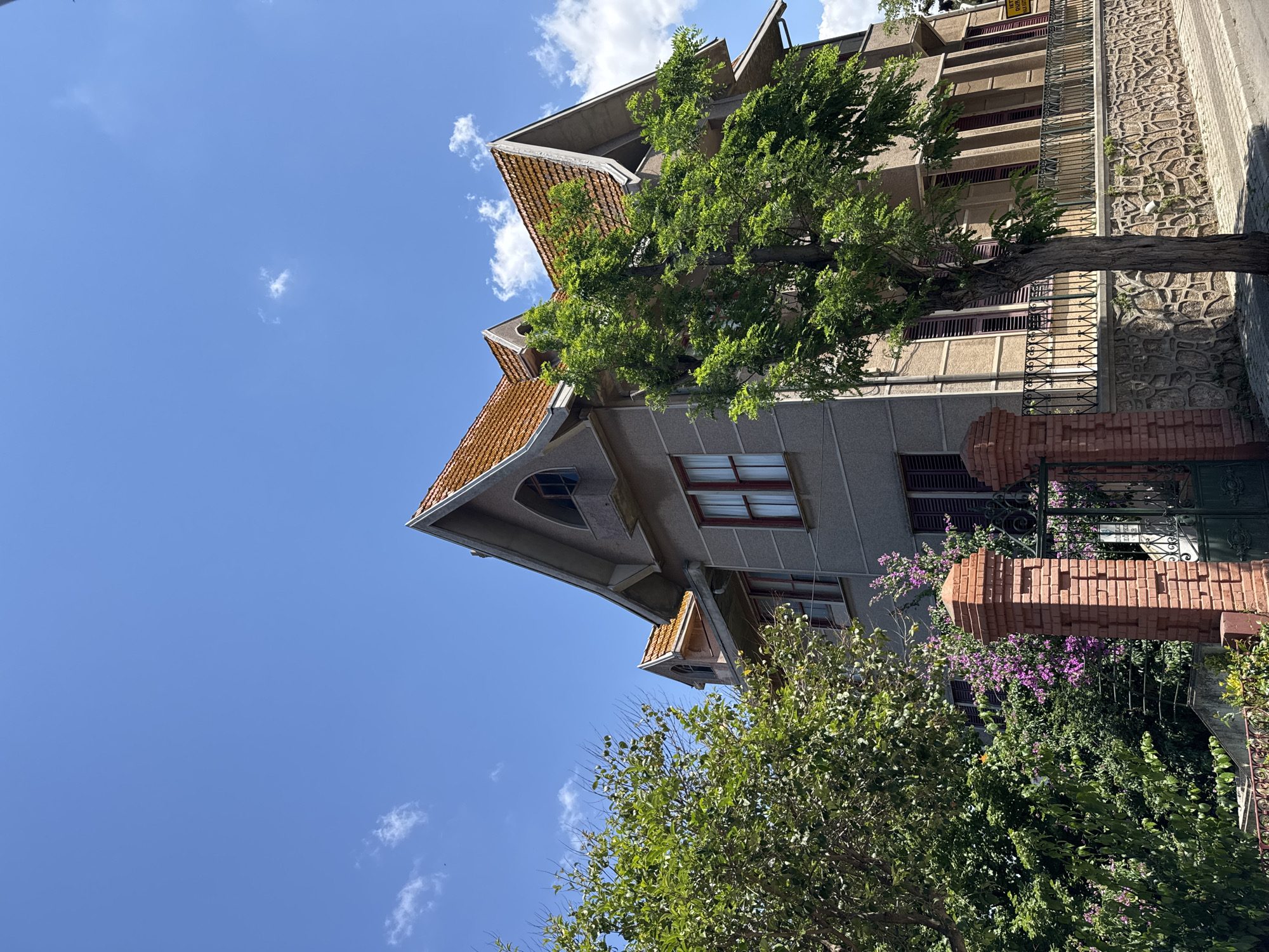 The image captures a two-story building with a grey facade and a distinctive orange tiled roof, situated in the city of Prince Islands, Turkey. The architecture features sharp gables and arched windows on the upper level. In the foreground, a mature tree with vibrant green foliage stands prominently, partially obscuring the house. To the left of the house, dense green bushes and flowering purple plants add a touch of color. A brick gatepost with a wrought-iron gate and fence lines the property, with a stone wall forming the lower boundary. The scene is bathed in sunlight under a clear blue sky dotted with a few white clouds, suggesting a pleasant, daytime setting. No people are visible in the frame.