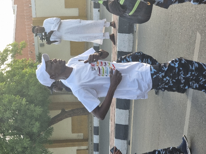 The image shows a man in a white t-shirt and a white cap running down a street in Maiduguri, Nigeria. He is wearing blue and black camouflage pants. He is running with his right arm bent, and he appears to be holding something with his left hand. Behind him, a person in camouflage pants walks, and another man wearing a white robe and a white cap appears to be filming the scene. In the background, a building with a yellow wall and a black fence is visible. The scene is a sunny day, and the street is paved with black asphalt and bordered by a black and white curb. A tree is to the left of the image. The coordinates provided indicate the location is in Maiduguri, Nigeria.