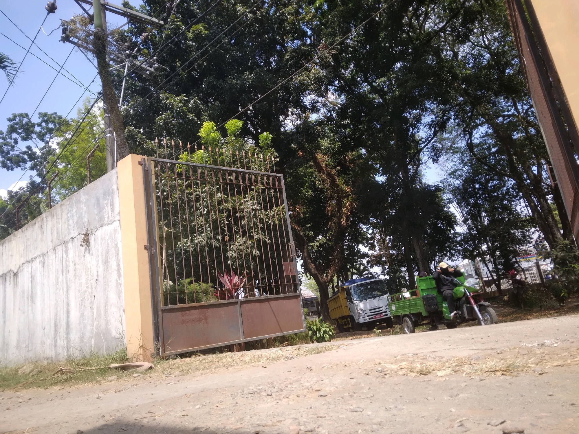 The image depicts an outdoor scene in Desa Pamijen, Kecamatan Sokaraja, Indonesia, with a focus on a rusty metal gate and a concrete wall. Behind the gate, lush green foliage and trees are visible, suggesting a natural or well-maintained area. In the background, a small, light-colored truck is parked next to a green, three-wheeled vehicle with a rider. The ground in the foreground is unpaved, with dirt and gravel indicating a rural or less developed setting. Overhead, a utility pole with electrical wires crosses the frame, a common sight in Indonesian towns and villages. The lighting suggests it is daytime, with shadows indicating the sun's position. The overall impression is one of everyday life in a semi-urban or rural Indonesian location.