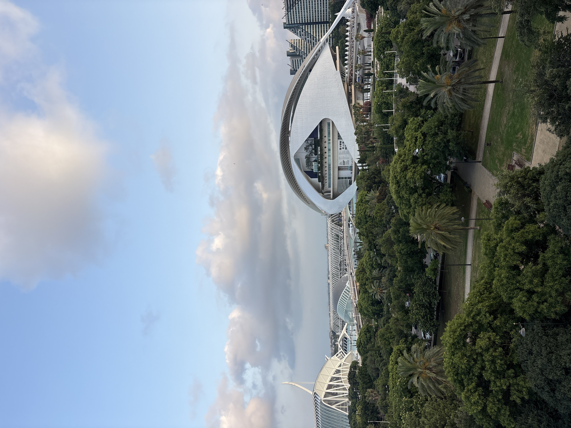 This is an outdoor, eye-level, full shot of the City of Arts and Sciences in Valencia, Spain. The architecture features striking white, futuristic buildings. The primary subject is the Hemisfèric, a building designed to resemble a giant eye, with its distinctive curved structure and the eye-shaped opening. In the foreground, lush green trees and palm trees line pathways and green spaces, indicating a park-like setting. Beyond the Hemisfèric, other structures of the City of Arts and Sciences are visible, including what appears to be the Palau de les Arts Reina Sofía. The sky is partly cloudy with patches of blue, suggesting it is daytime, likely late afternoon or early evening based on the soft lighting. The scene evokes a sense of modern architectural wonder and tranquility within a green urban environment. No people are prominently visible.