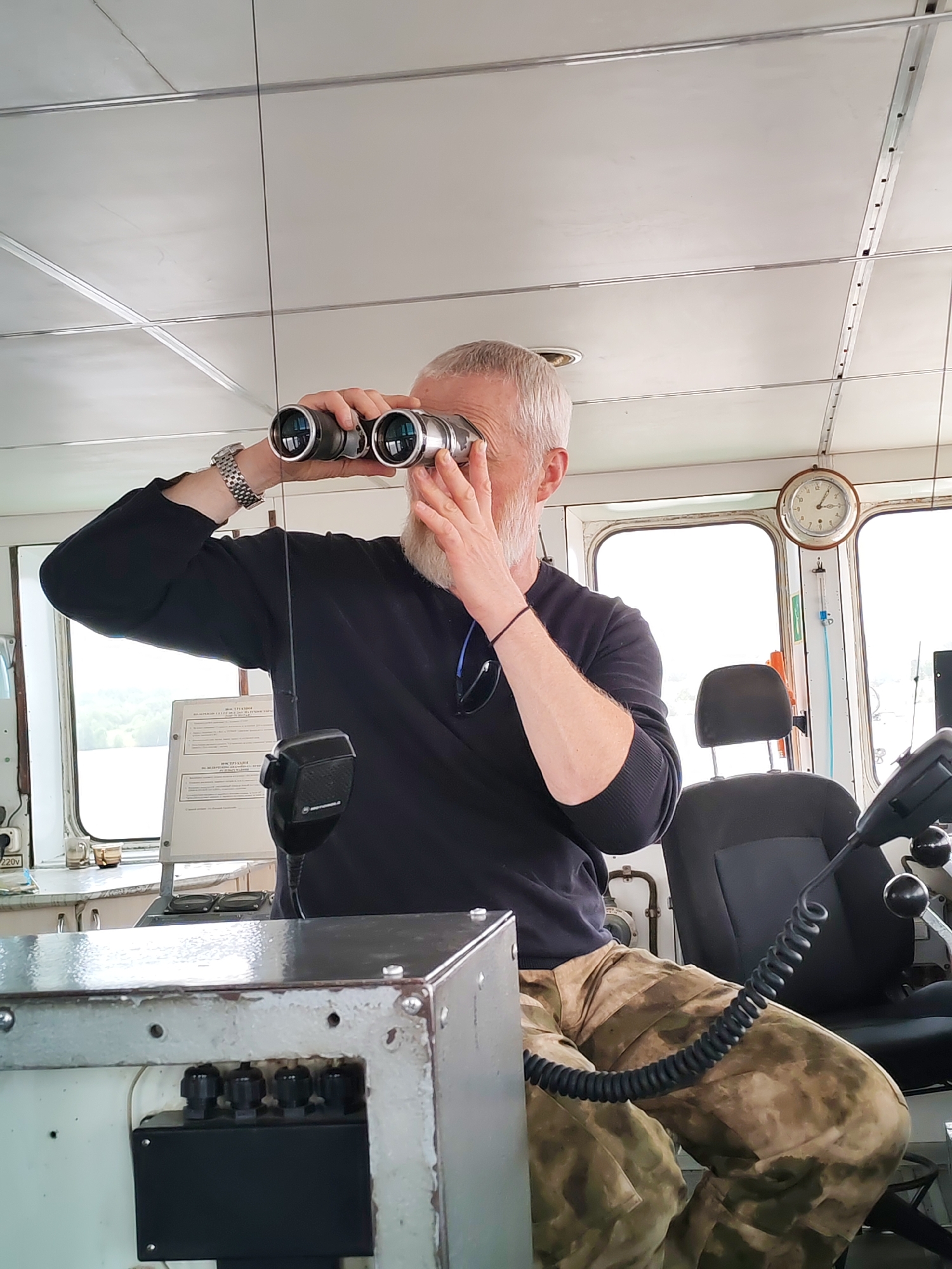 A middle-aged man with white hair and a beard is standing inside what appears to be the bridge of a boat or ship. He is holding binoculars to his eyes, looking out of a window. He is wearing a dark blue long-sleeved shirt and camouflage pants. A silver watch is visible on his left wrist. A microphone hangs in front of him. Behind him is a swivel chair, a clock on the wall, and a window showing a view of a riverbank. A metal control box with various knobs and switches sits in the foreground. The man's location is in Раменье, Russia.