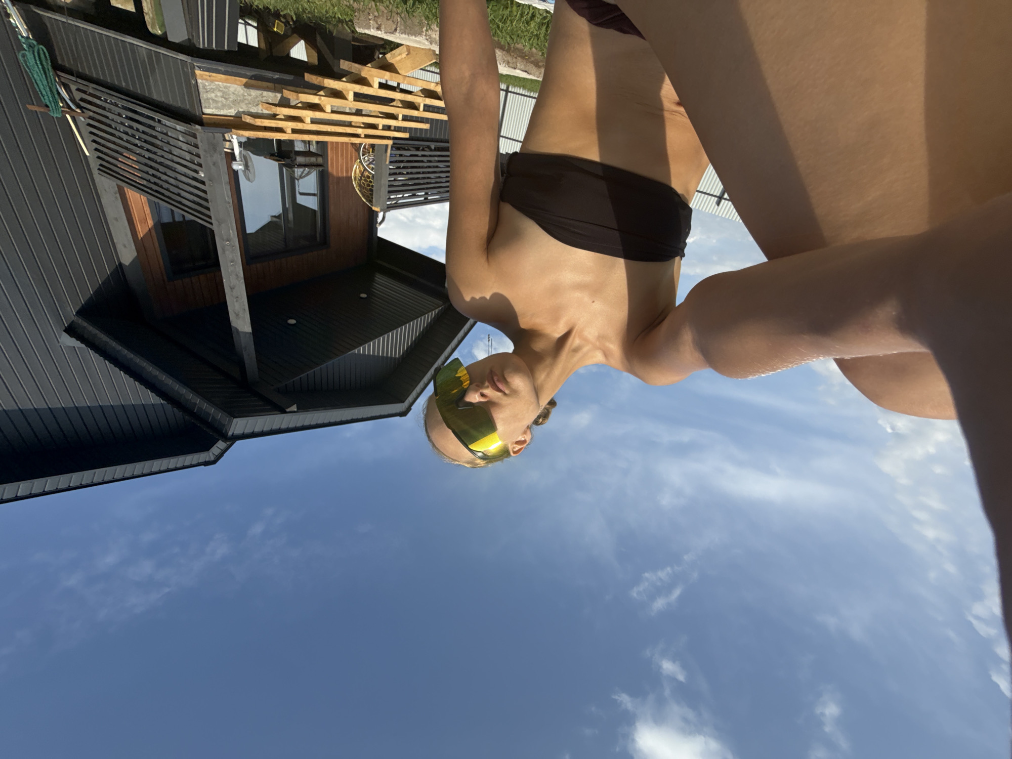A woman wearing sunglasses and a black bikini top is sitting outdoors on a sunny day. She is angled towards the camera, with her legs crossed. Behind her, a dark gray modern house with wooden accents and a balcony is visible. Wooden steps lead up to the balcony, and a green rope is draped over the railing. The sky is blue with scattered white clouds. The scene appears to be in a rural or suburban setting, possibly in or near the village of Podshivalovo, Russia, given the presence of a modern house and outdoor recreational space.