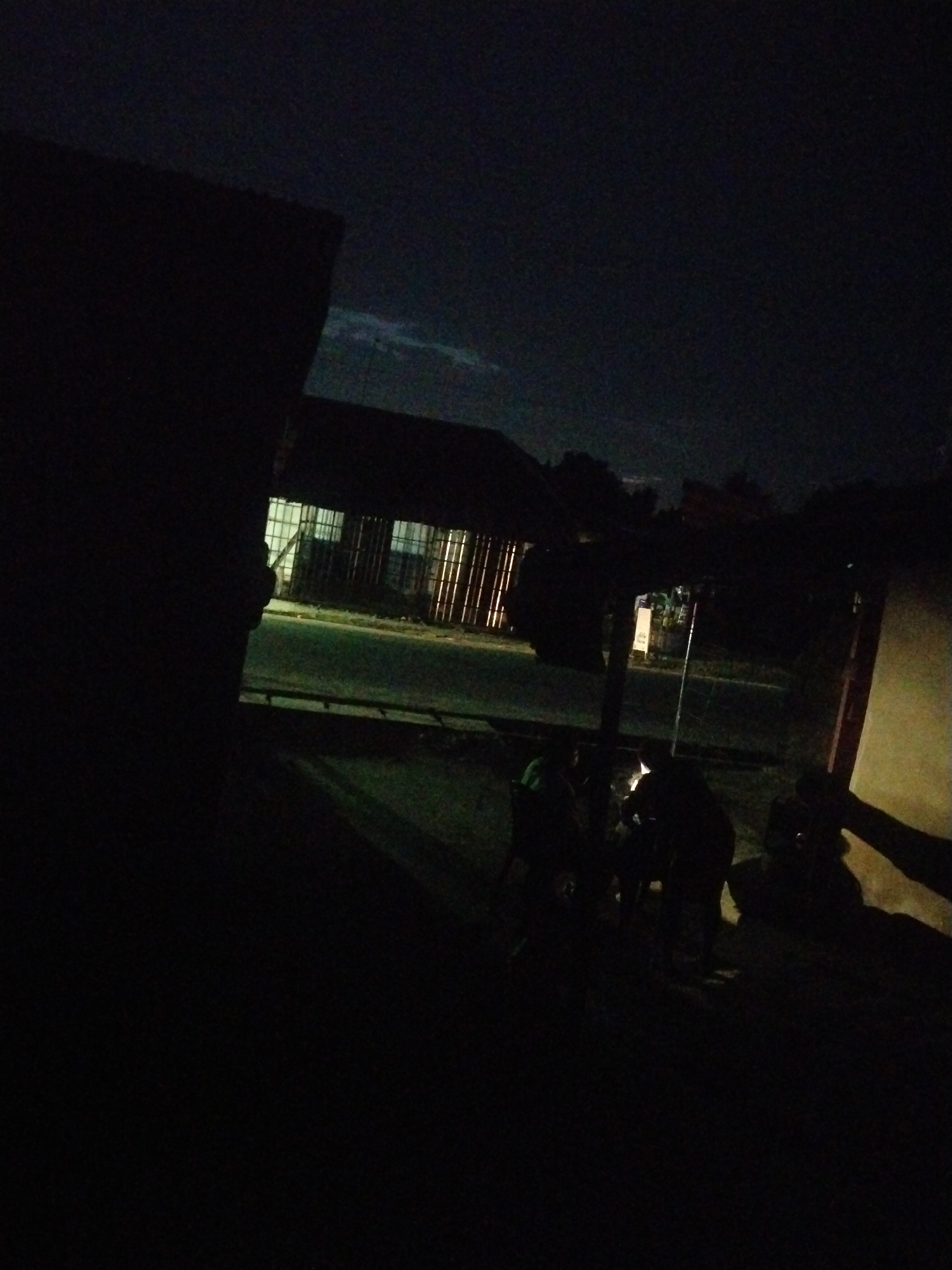 The image depicts a dark, nighttime scene in Uyo, Nigeria, with a building characterized by barred windows casting light onto a road. In the foreground, a group of individuals are gathered, some seated, with one person appearing to be holding a light source. The setting suggests an outdoor gathering or a scene at a roadside establishment.