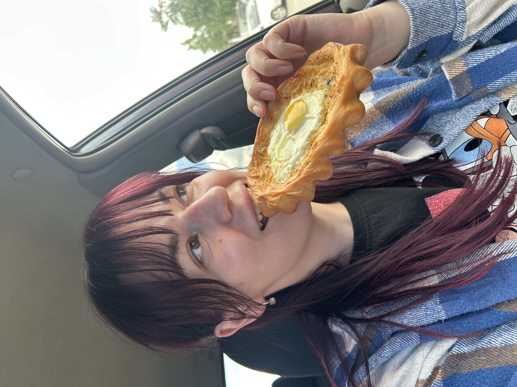 A woman with long, dark red hair and bangs, wearing a blue and white plaid shirt over a black t-shirt with a cartoon graphic, is seated in the driver's side of a car. She is holding a pastry, which appears to be a type of bread with an egg and cheese filling, in her right hand and is in the process of taking a bite. Her left hand is near her face, and she is looking off to the right. The interior of the car, including the dashboard and door panel, is visible. The background shows a blurry outdoor scene with trees and a white car parked. The location is specified as Chaikovsky, Russia.