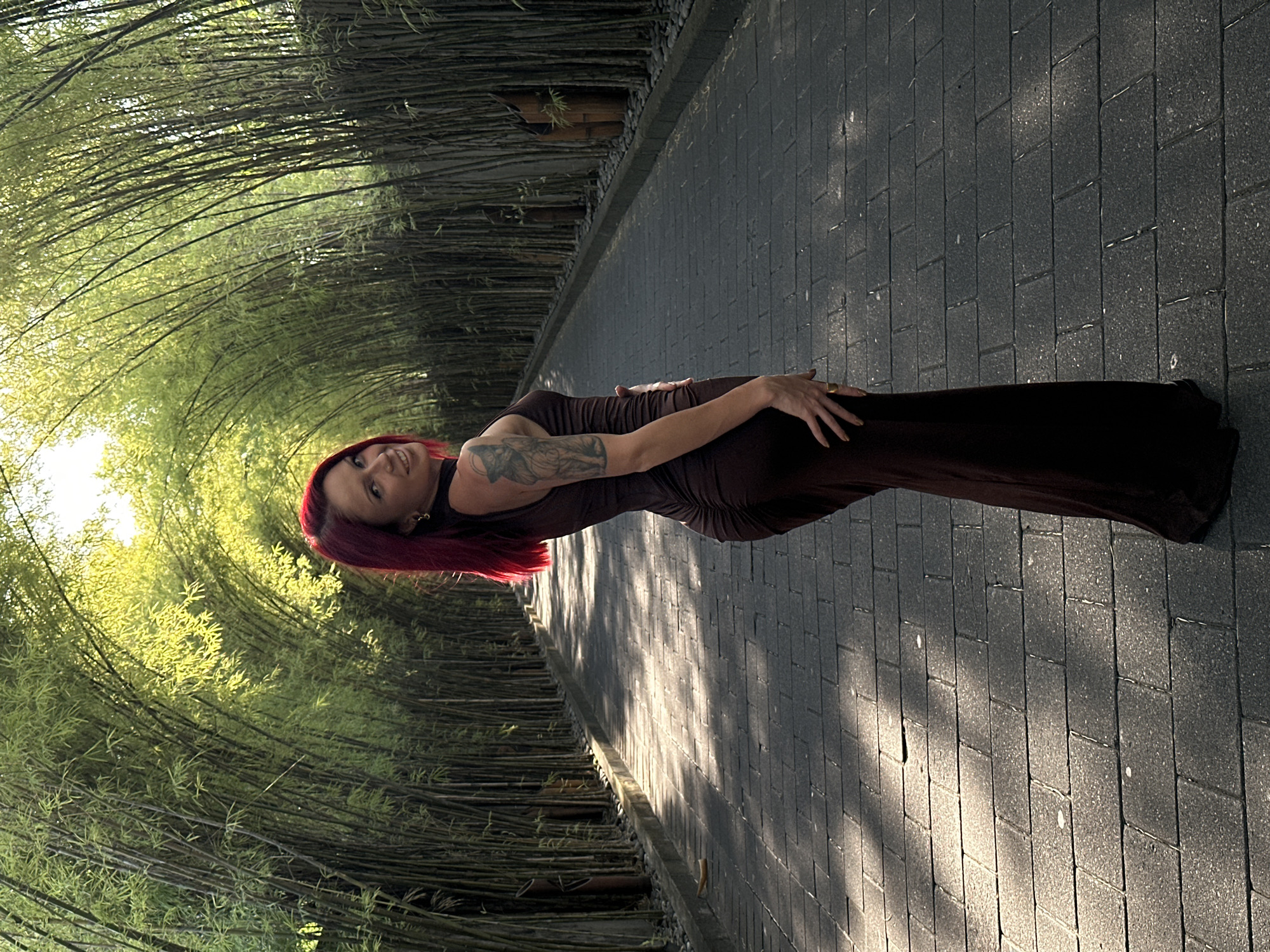 A woman with red hair, wearing a dark brown form-fitting dress, is posing on a paved walkway lined with bamboo trees, possibly in North Kuta, Indonesia. The walkway is made of dark rectangular paving stones, with a lighter, sunlit area on the right. The bamboo trees arch overhead, creating a tunnel-like effect, with bright sunlight filtering through the leaves. The woman is facing towards the viewer with her arm across her body. A tattoo is visible on her upper arm. The overall lighting suggests it's daytime.