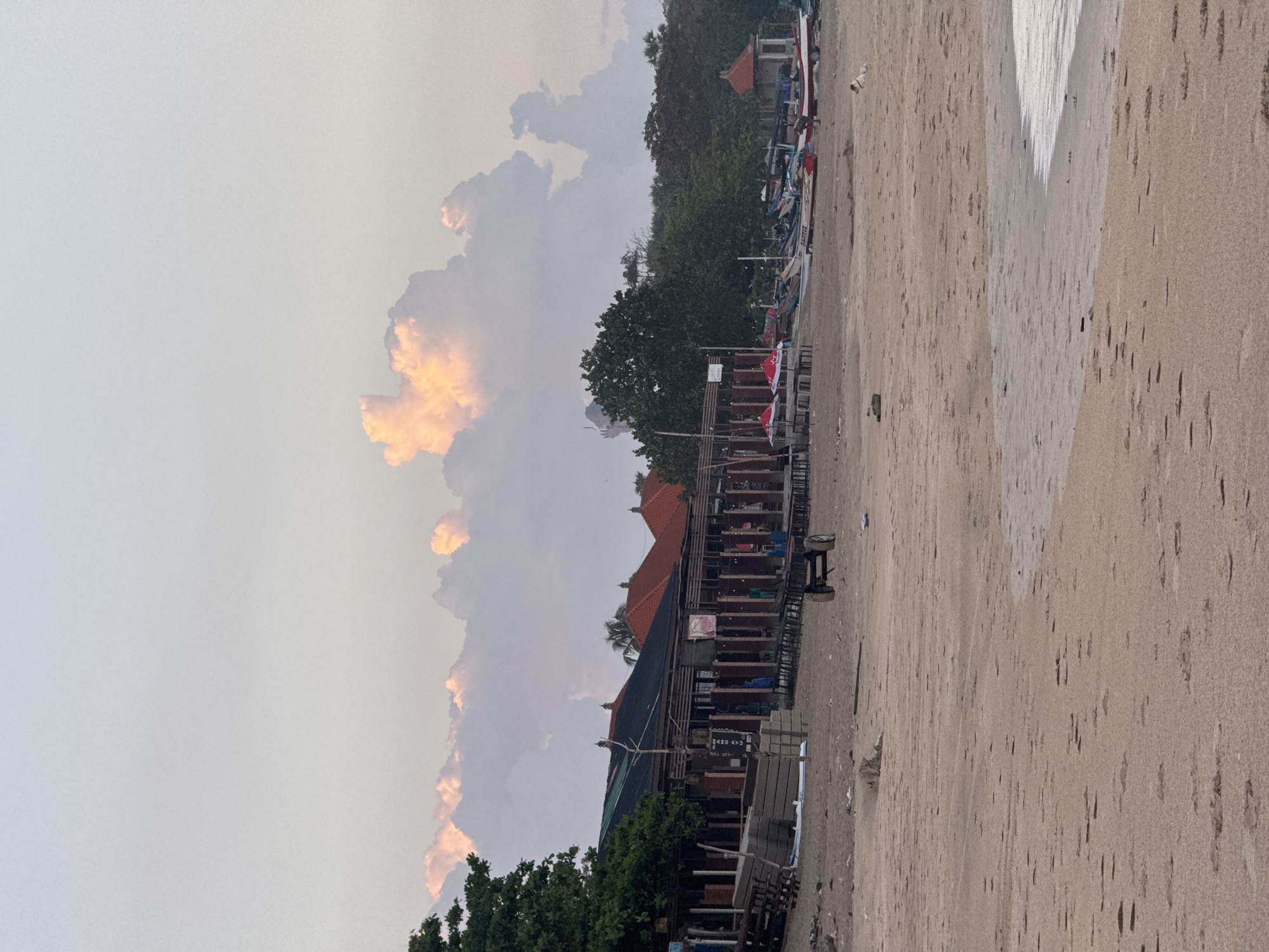 The image depicts a beach scene in Kuta, Indonesia. The foreground features a sandy beach with the tide line visible. The middle ground shows several buildings with dark roofs and exposed wooden structures, possibly beachside restaurants or shops. Trees are interspersed among the buildings. In the background, there is a row of parked fishing boats and more lush vegetation. The sky is overcast with large, cumulus clouds tinged with orange light, suggesting either sunrise or sunset. No people are clearly visible in the image.