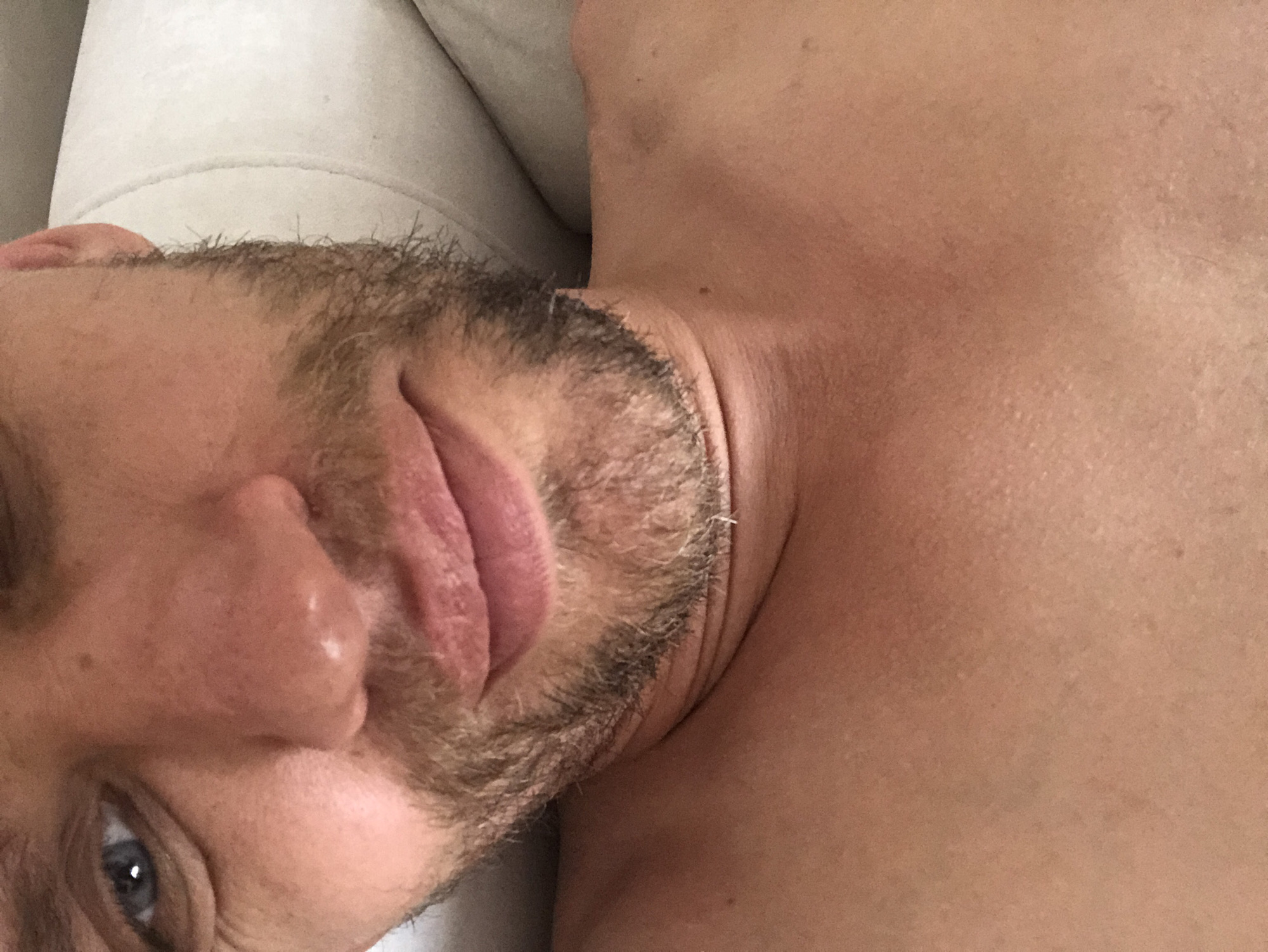 The image is a close-up, overhead shot of a fair-skinned individual lying on their back. The person has blue eyes, a light brown stubble beard, and is shirtless, revealing their chest and neck. The background shows a white, plush surface, possibly a pillow or couch, with a slight shadow indicating a light source from the right. The scene is indoors, with no discernible urban or city features visible. The person appears relaxed, with their head tilted slightly.
