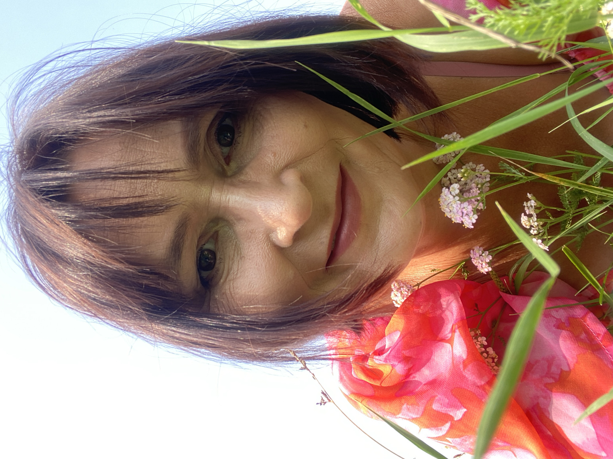 An extreme close-up shot captures a woman's face as she looks down at the camera, surrounded by tall green grass and small pink wildflowers. Her brown hair is styled with bangs that partially obscure her forehead. She wears a bright pink and orange floral patterned scarf draped around her shoulders, partially obscuring a pink garment underneath. The background is a bright, clear sky. The image is an outdoor shot and does not contain any elements indicating a specific location in Novokuznetsk, Russia.