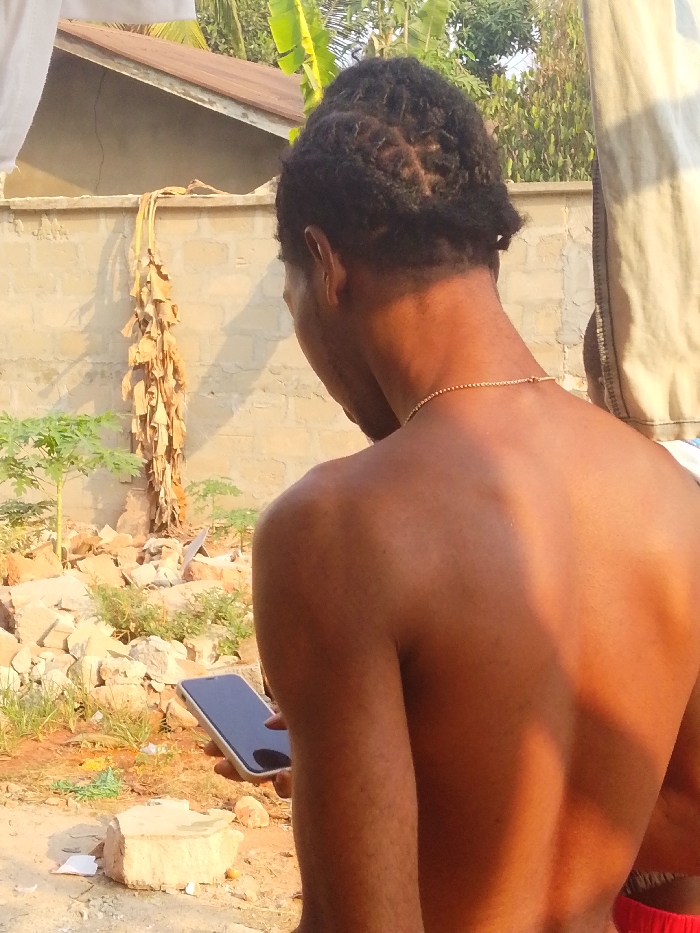 In Enugwu-Ukwu, Nigeria, an outdoor scene shows an individual from behind, primarily focusing on their upper torso and head. The person has dark skin, short, dark dreadlocks or twists, and wears a thin gold-colored chain around their neck. Their upper body is bare, and a red garment is partially visible at the bottom right. The individual holds a smartphone with a black screen and a light-colored frame vertically in their left hand, appearing to be focused on the device.
The background features a light-colored concrete block wall. Above the wall on the left, a portion of a brown corrugated metal roof is visible. Dried, brown banana leaves hang against the wall. The ground in the mid-ground is uneven, covered with loose soil, scattered concrete blocks, and rocks, interspersed with small green leafy plants, possibly young papaya trees. Beyond the wall, dense green foliage suggests further vegetation. The scene is brightly lit by direct sunlight, casting shadows on the wall and the subject's back.