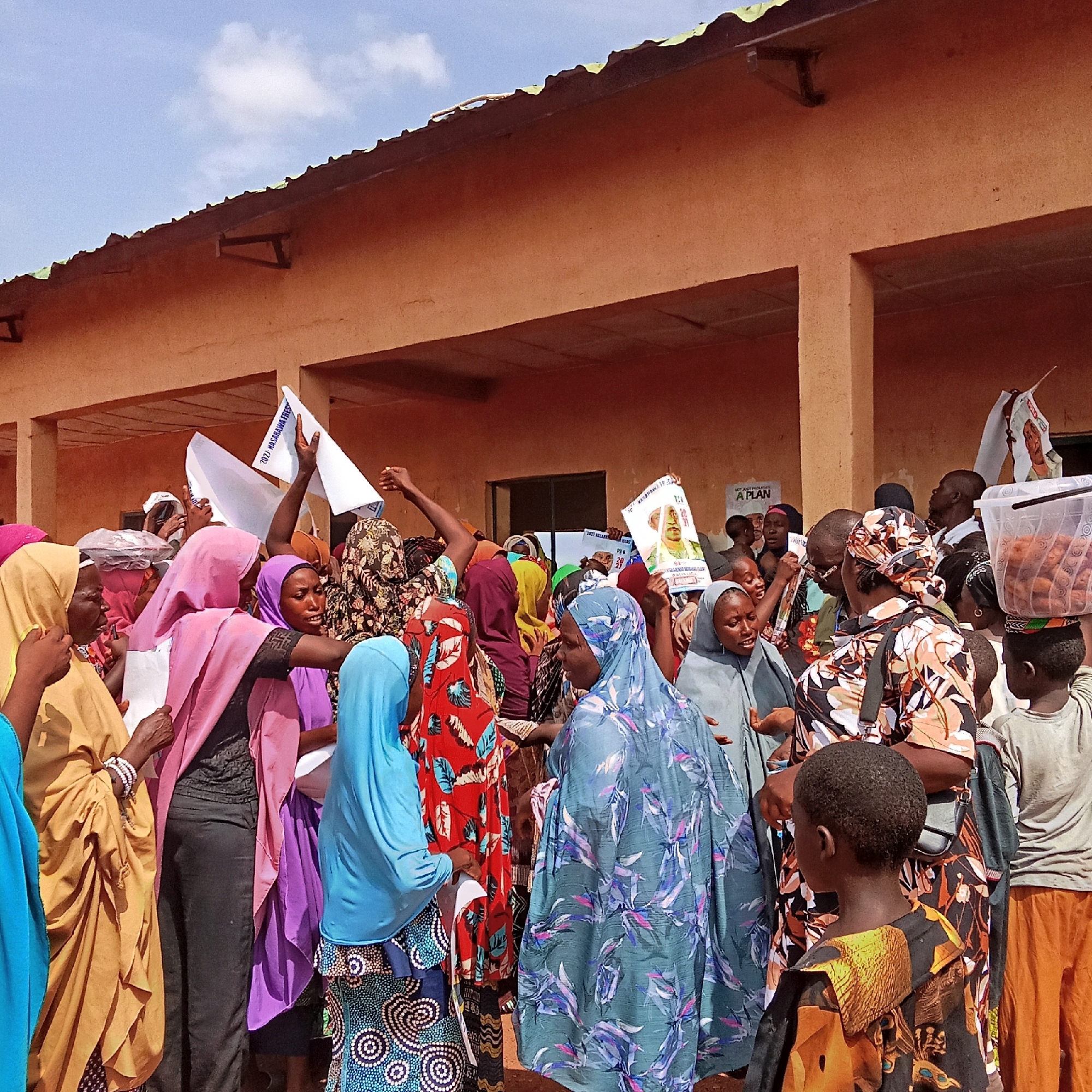 The image shows a crowd of people, many wearing traditional clothing and head coverings, gathered in front of a light-orange building. Some individuals hold signs with images and text, possibly related to a political campaign. A child is carrying a container of what appear to be baked goods on their head. The sky is visible in the background, with a few clouds. The image was taken in Akwanga, Nigeria, as printed on a sign.
