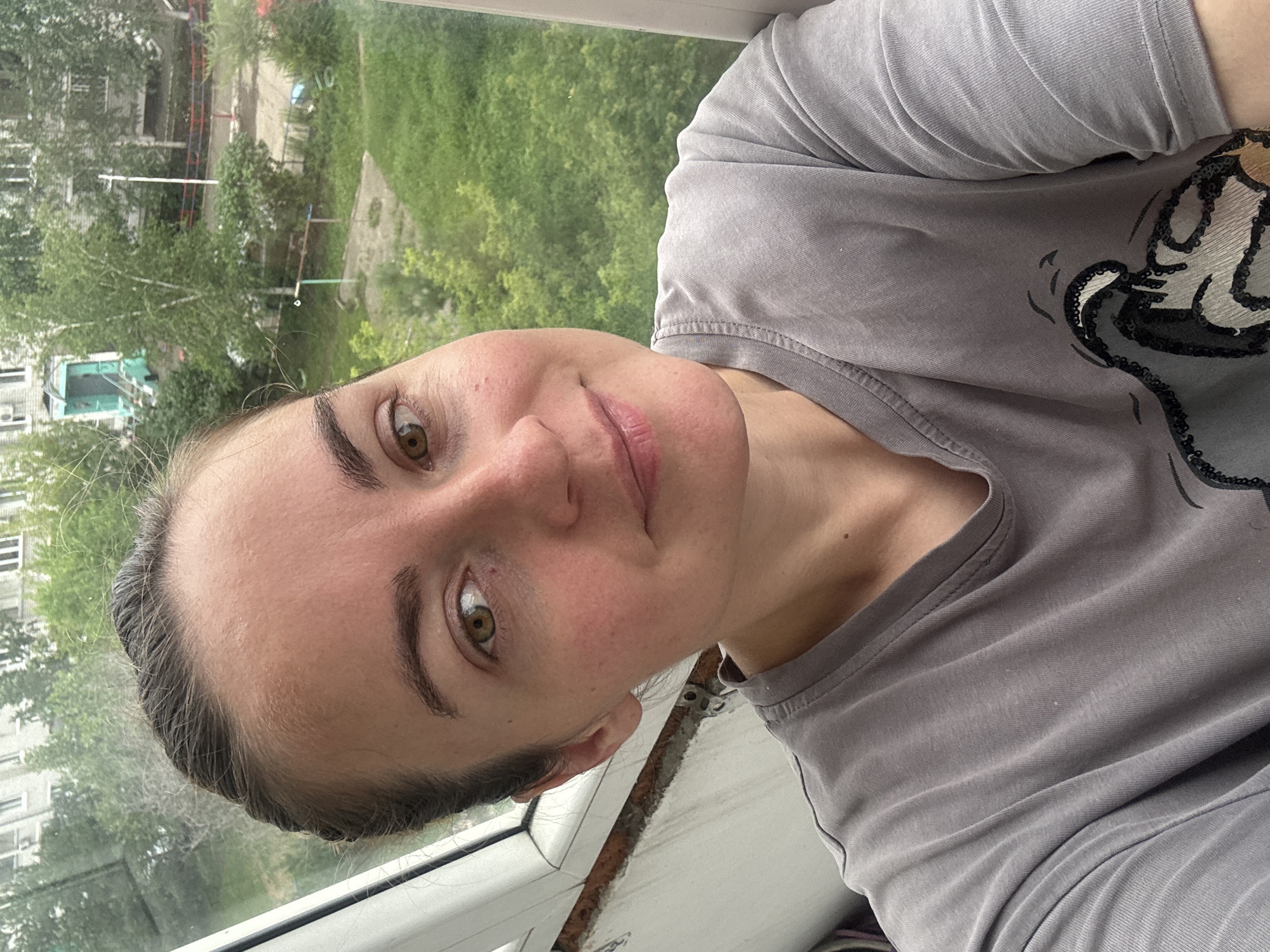 A close-up, shoulder-up selfie of a woman with brown hair pulled back, looking slightly off-camera and smiling gently. She is wearing a grey t-shirt with a graphic print of a cartoon character. The background suggests an urban residential setting with apartment buildings and green trees visible through a window. The environment appears to be Blagoveshchensk, Russia, possibly taken from a balcony or apartment window overlooking a courtyard. The lighting is overcast, indicating it's likely daytime.