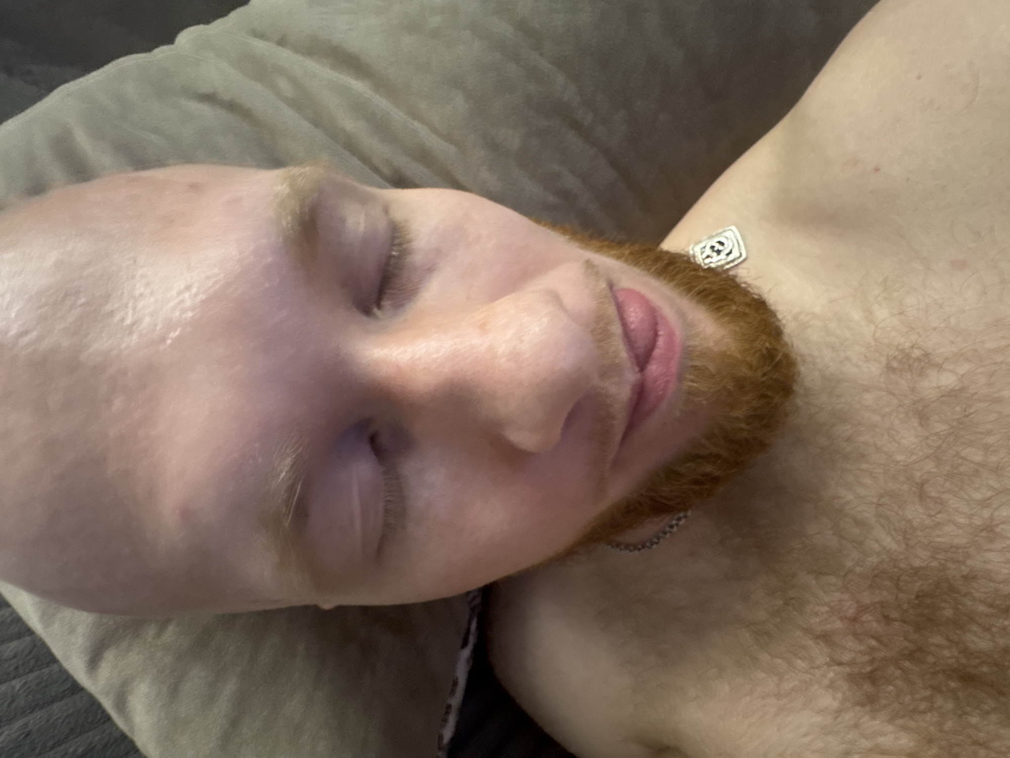 A close-up, top-down shot depicts a person lying down with their eyes closed. The individual has a bald head and a red beard. Their skin tone is light. They are wearing a silver necklace. The person is lying on a gray pillow. The photo's location is not discernible from the image.
