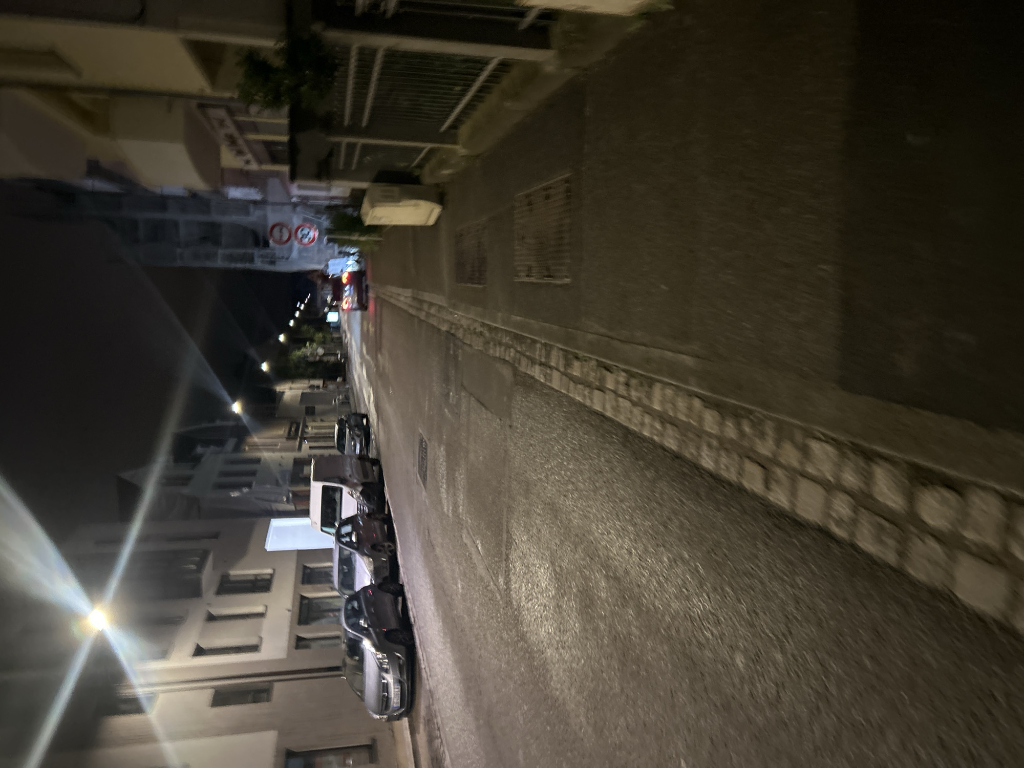 The image depicts a street at night in Strasbourg, France. The street is lined with parked cars on the left side. Buildings are visible on both sides of the street, illuminated by streetlights. Traffic signs are also visible in the distance. The road surface appears wet.