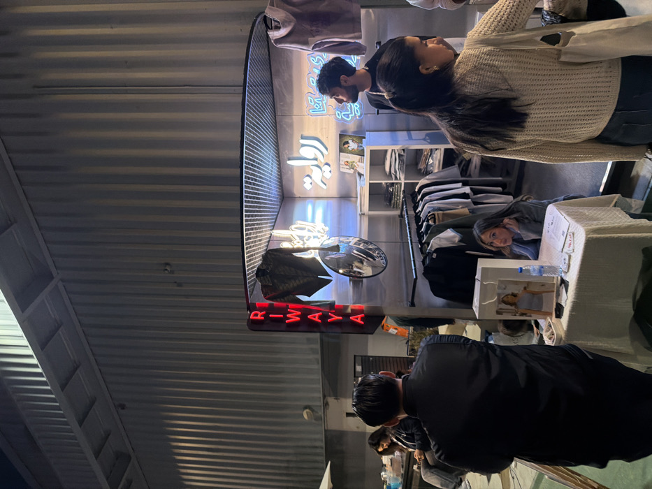 A group of people gathered outside a store with a neon sign reading "RIWAYA." Some are browsing clothes on racks, while others are standing near a display table. A woman is leaning over the table, and a man in a black coat is looking at something below. The store has bright neon lights and reflective surfaces, and the ceiling above is made of corrugated metal.