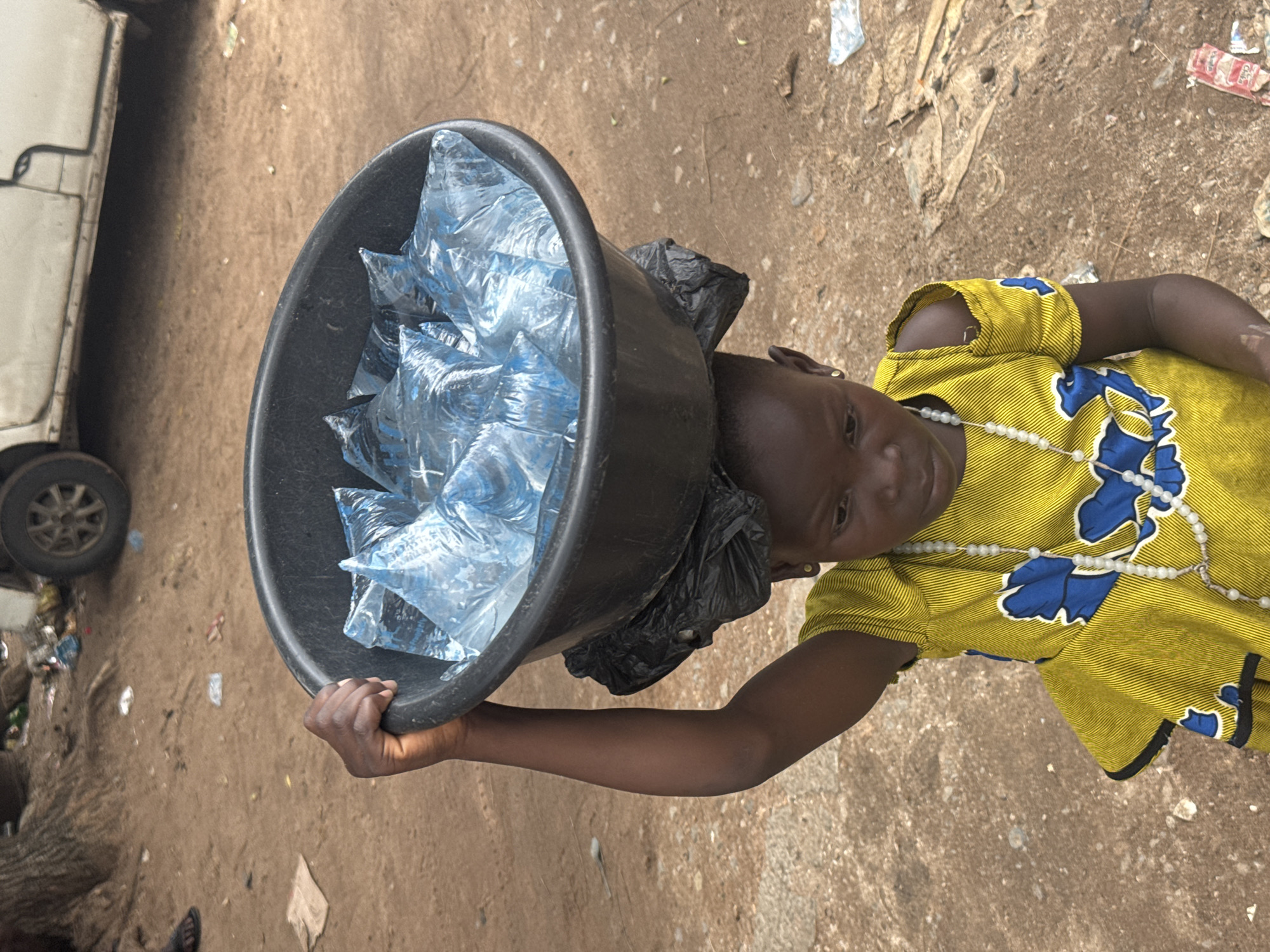 In the city of Owerri, Nigeria, a young girl, likely a street vendor, carries a large black basin filled with plastic sachets of water on her head. She is wearing a bright yellow dress adorned with blue floral patterns and a beaded necklace. The girl is looking down, her expression serious as she balances the heavy load. The ground around her is dirt and strewn with debris, including discarded plastic bags and wrappers, suggesting an outdoor market or a street environment. In the background, a white vehicle is partially visible, along with tree roots and more scattered litter. The scene depicts a common sight of child labor and the challenges of daily life in this urban setting.