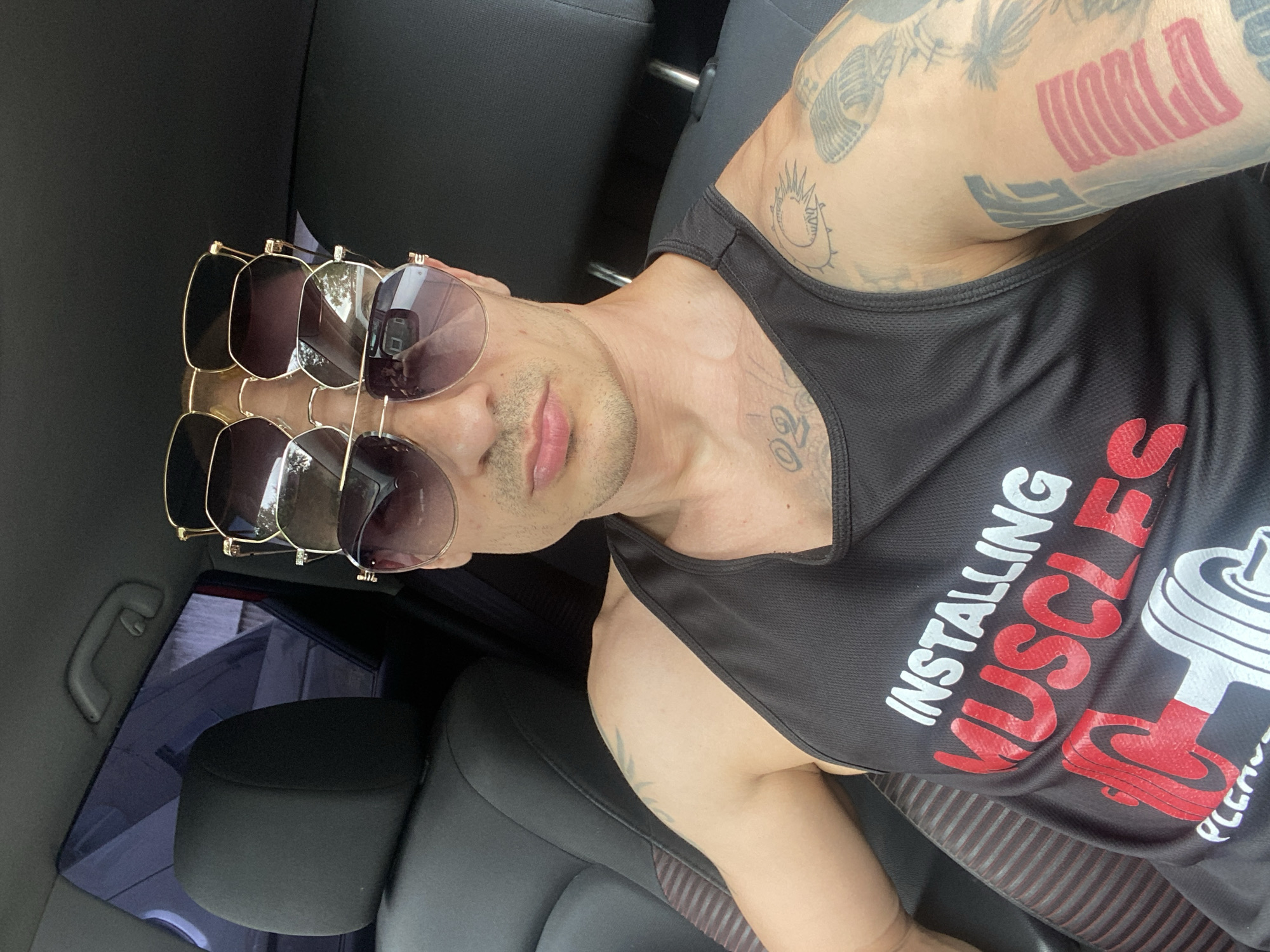 A man is in a car, wearing a black tank top with "INSTALLING MUSCLES PLEASE WAIT" printed on it and has several pairs of sunglasses stacked on his head. His arms are visible, and are covered in tattoos. The man is located in a car, with black leather seats. There is no evidence to suggest the man is located in the city of Pleven, Bulgaria.