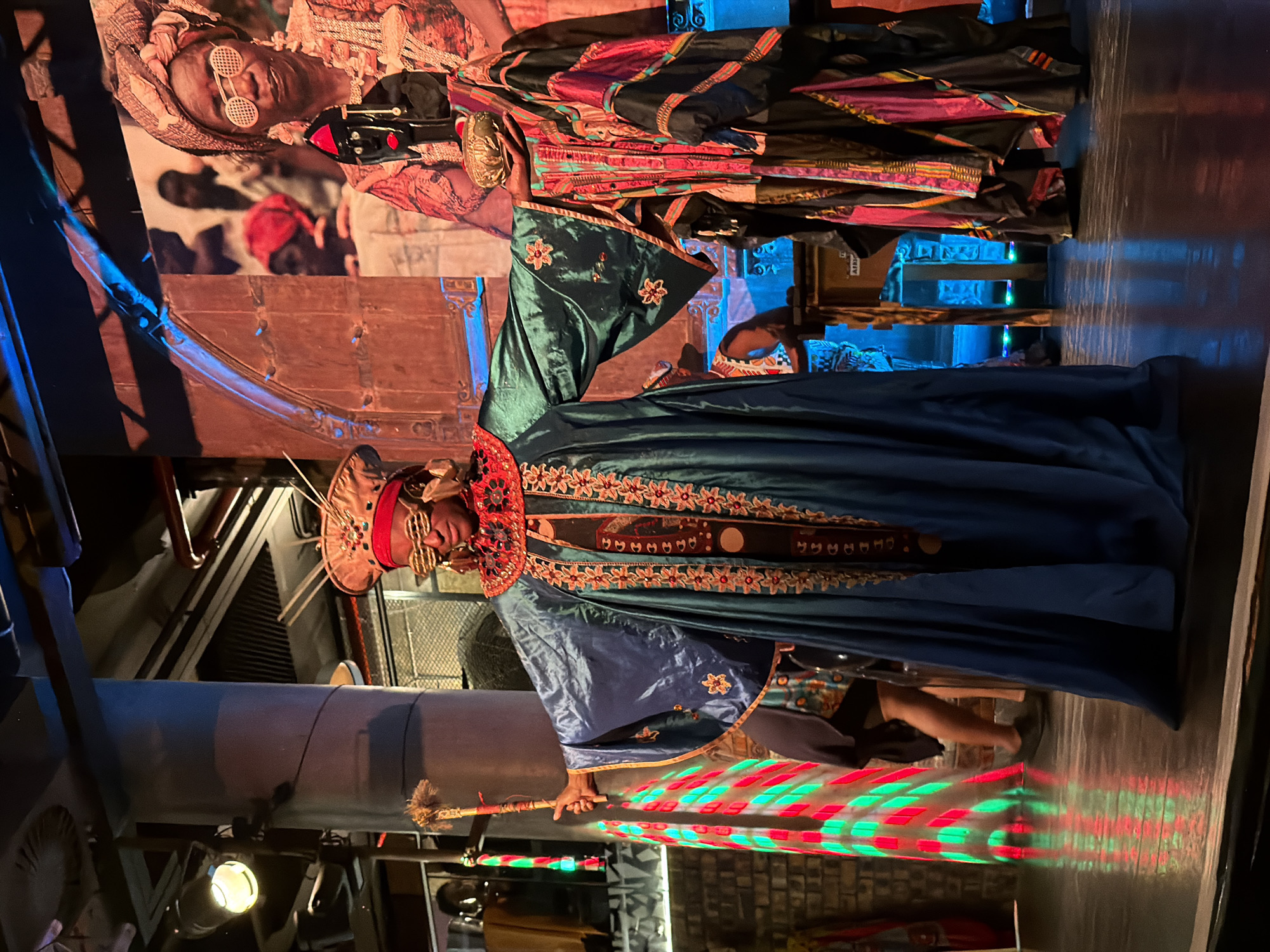 This image captures a person in an elaborate, culturally inspired costume within a dimly lit, theatrical indoor setting, likely in Cape Town, South Africa. The central figure wears a rich, deep blue-green robe adorned with intricate red and gold embroidery along the collar and front opening. A matching broad-brimmed hat or headpiece with similar embellishments covers their head, and they wear distinctive round, white-rimmed sunglasses.
The individual holds a decorated staff or scepter upright in their right hand. The staff's lower portion reflects dynamic red and green diamond-patterned lights onto the dark floor, suggesting an active light display, possibly from a projector or stage lighting.
In the background, a large vertical image, possibly a banner or screen, displays another person in traditional attire and round sunglasses, mirroring the style of the main subject. This backdrop, along with ornate wooden structures and soft blue ambient lighting, creates a vibrant and culturally rich atmosphere. Overhead, industrial pipes and vents are visible, blending with the decorative elements.
The overall scene suggests a cultural performance, an exhibition, or a themed event celebrating African heritage. The artificial lighting indicates an evening or indoor occasion. Faint text "AFRICA" can be seen on a box in the background, reinforcing the cultural context of the setting.