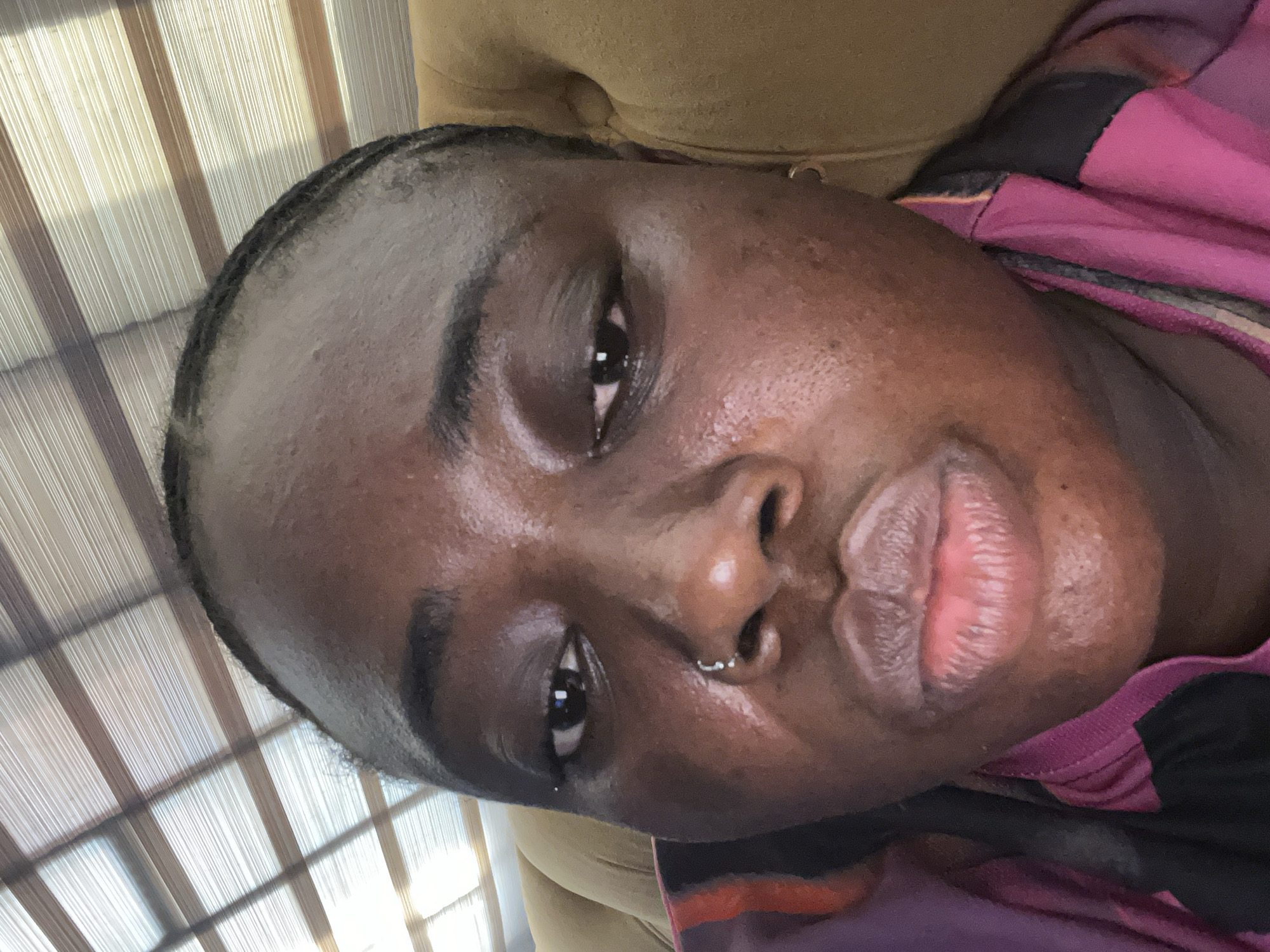 A close-up, rotated portrait of a person with dark skin. The person has dark eyes, dark eyebrows, and a nose piercing. They wear a pink and black garment. The background shows window blinds and a tan-colored surface. Location is unspecified in the image and cannot be determined to be Kubwa, Nigeria.