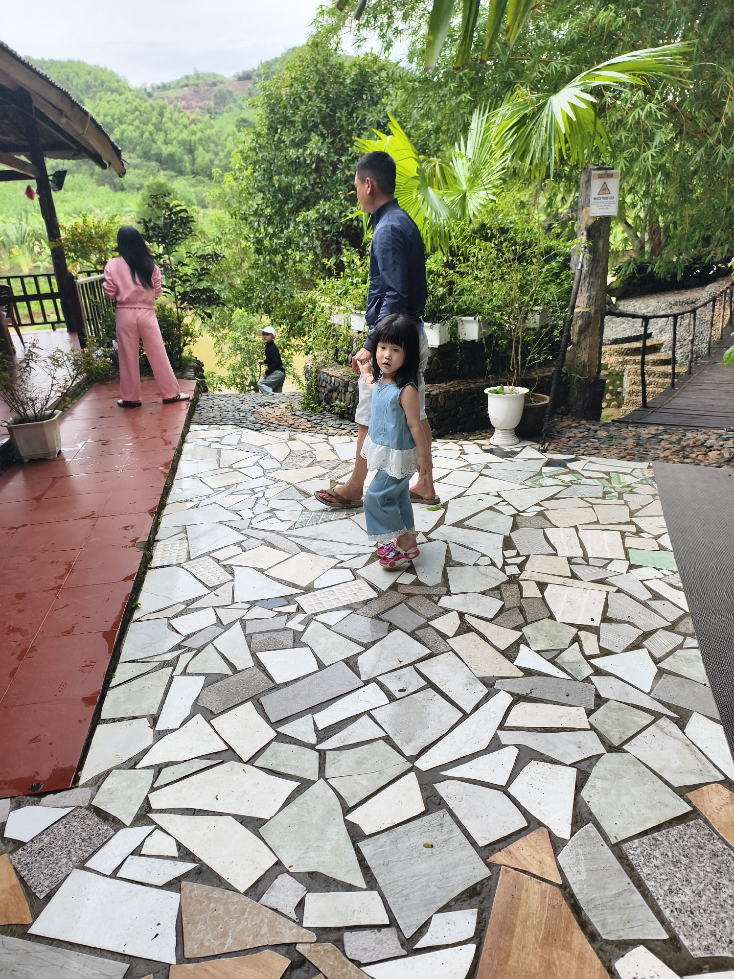 The image depicts an outdoor scene in an environment with lush greenery. A young child, dressed in a blue denim outfit and pink shoes, stands on a tiled walkway with a geometric mosaic pattern. An adult male stands behind the child, dressed in a dark blue shirt and light-colored shorts. Another person in a pink pantsuit is visible in the background, near the edge of a covered patio with red tile flooring. In the distance, a person in dark clothing walks down a set of stairs leading to the river. A dense green forest rises up behind them and there is a sign that says "Caution". The location appears to be Unknown, Unknown.