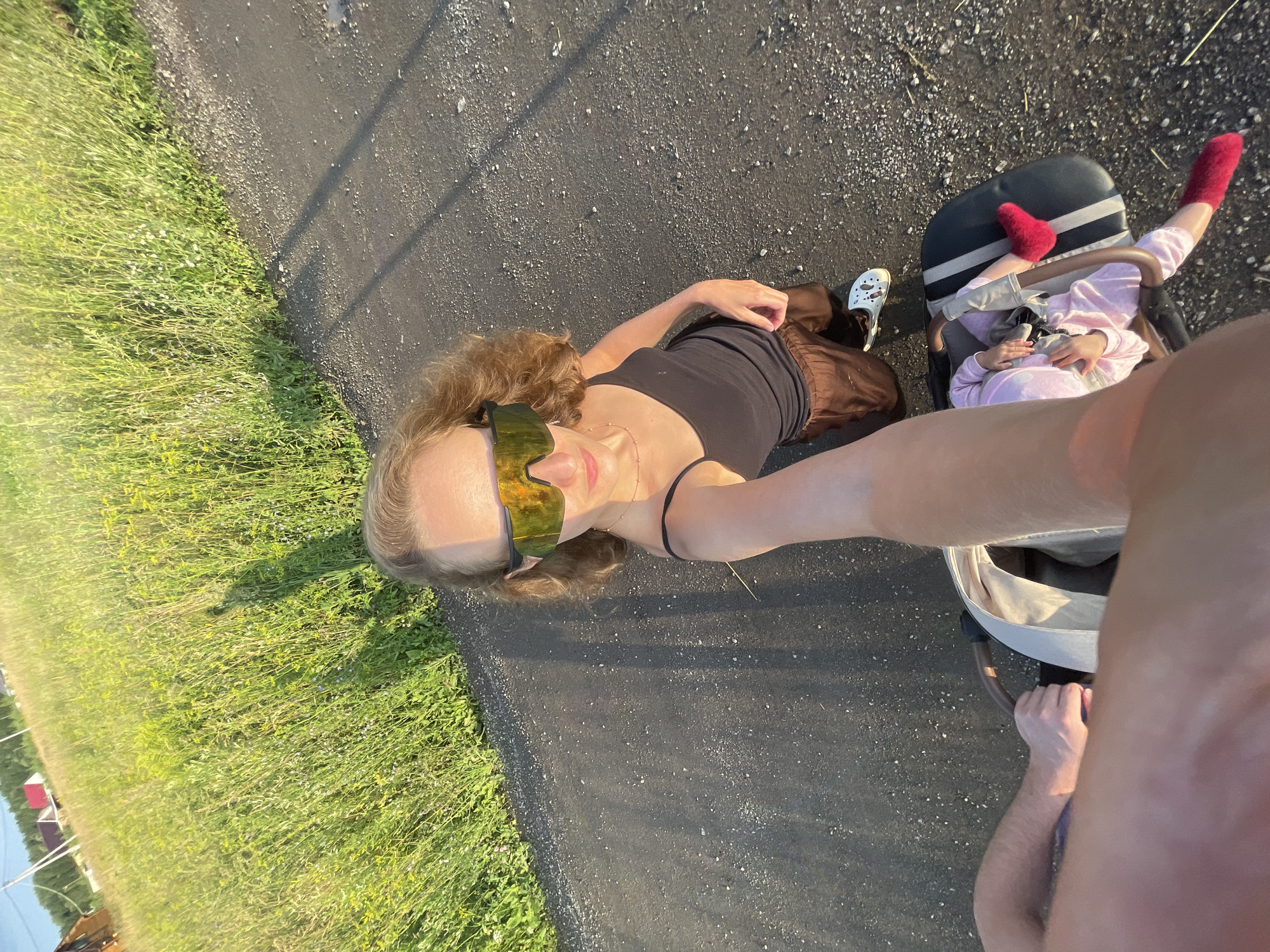 A woman wearing yellow reflective sunglasses, a black tank top, and brown pants is taking a selfie while kneeling next to a stroller on a paved path. A baby in a pink outfit is in the stroller, wearing red socks. The woman's right arm is extended towards the camera, holding a thin stick. A person's arm and shoulder are visible in the foreground. To the left of the path is a grassy field with wildflowers. In the background, houses and trees are visible under a clear sky. The scene appears to be in the village of Podshivalovo, Russia.