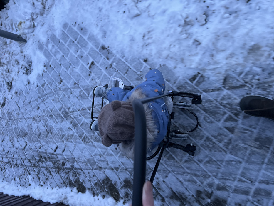 This image, captured in Khimki, Russia, presents a top-down view of a winter outdoor scene. A single child is visible, seated in a black metal sled-stroller that features both runners and wheels. The child wears a blue winter coat with a fur-trimmed hood, a brown hat with ear-like protrusions, and light-colored boots. The sled-stroller is being propelled forward, indicated by motion blur on the ground. A human hand, holding a black object (likely a mobile device), extends into the frame from the upper left, and a dark-booted foot is partially visible in the lower center, suggesting the presence of an adult pushing the stroller. The ground is a paved path with a diagonal block pattern, uniformly covered by a thin layer of snow and ice. To the left, a dark, vertical structure, possibly a fence or wall, is partially obscured by snow. To the right, an accumulation of snow and a grey cylindrical pole are present. The lighting is diffused daylight.