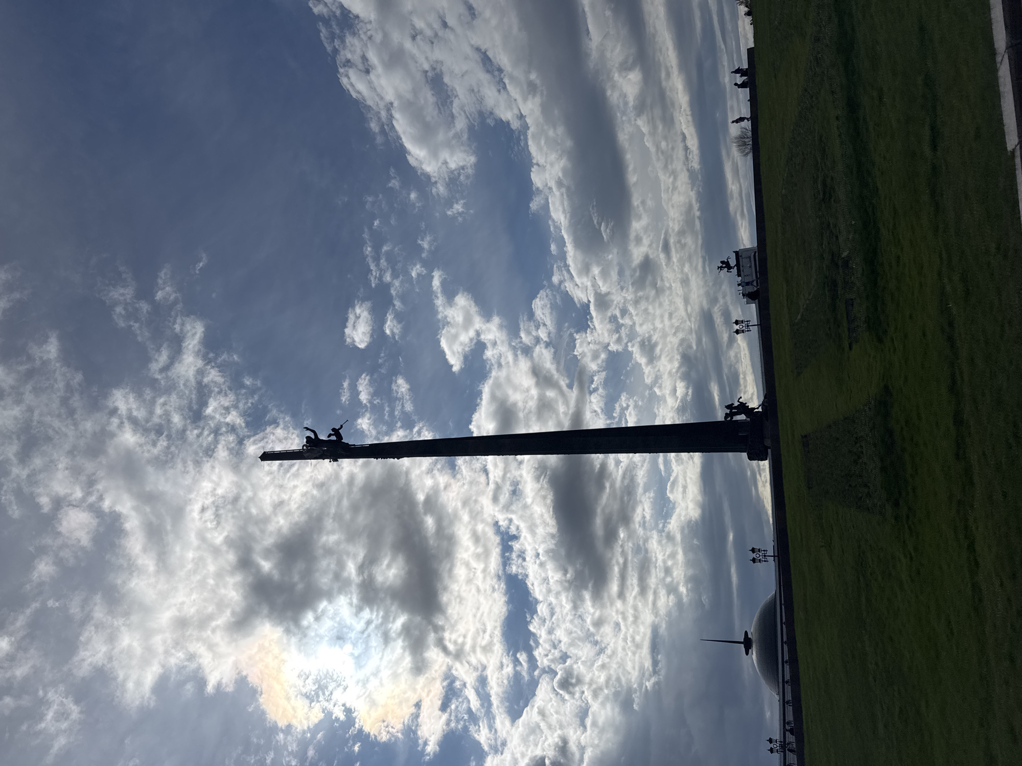 The image shows a tall, dark monument silhouetted against a cloudy sky in Moscow, Russia. At the top of the monument, figures of people are visible. Below, there is a green expanse of grass and, in the distance, a dome-shaped building with a spire. The monument is located in Victory Park, commemorating victory in the Great Patriotic War. Several objects are lined up on both sides of the monument.