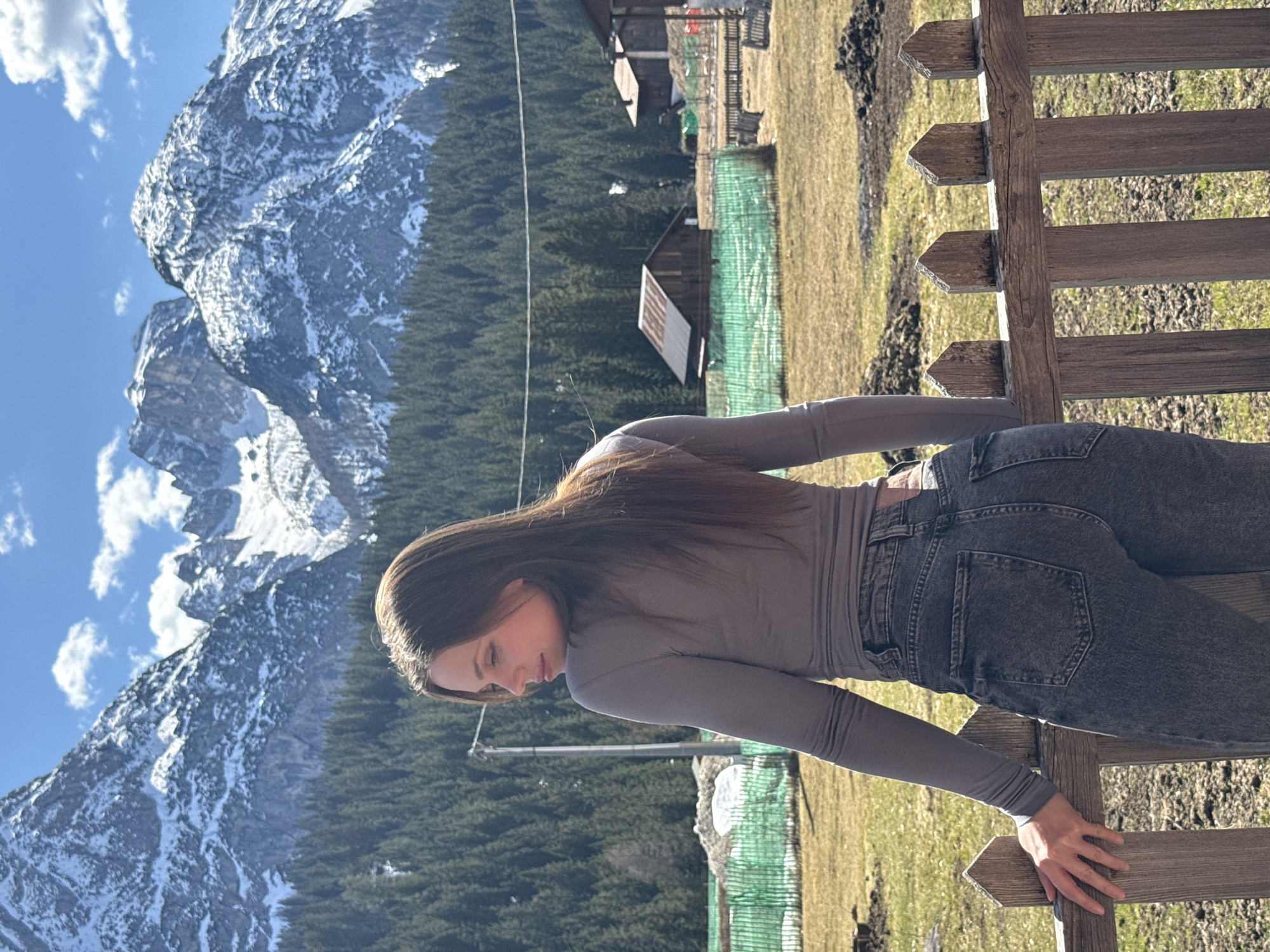 A woman with long brown hair is leaning on a wooden fence in Sappada, Italy. She is wearing a grey long-sleeved shirt and dark jeans. In the background, there is a mountain range covered in snow, a forest of pine trees, and several small wooden buildings. The sky is blue with scattered clouds.