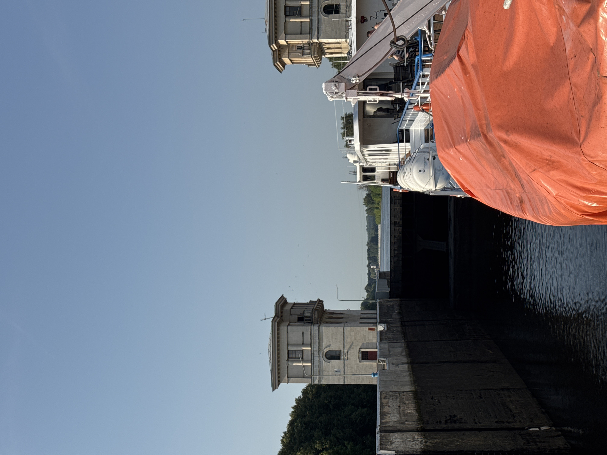 This is a vertical, eye-level, medium shot of a ship in a canal lock in Iksha, Russia. The sky is clear and blue. On the left is a concrete wall of the lock, and on the right is a white ship with an orange tarp-covered object in the foreground. Behind the ship, another building can be seen. The ship is docked in the lock. The canal lock is a part of the Moscow Canal, a waterway that connects Moscow to the Volga River. The canal lock is a concrete structure with two large gates. The gates are closed, and the water level in the lock is lower than the water level outside the lock. The ship is a passenger ship, and it is docked in the lock. The ship has a white hull, and it has a superstructure with windows. The ship has a lifeboat on the deck, and it is covered with an orange tarp. The building behind the ship is a two-story building with a white facade and a red door. The building has a balcony on the second floor. The building appears to be an administrative building or a control tower for the lock. The trees are visible in the background of the image, and they are green and lush.