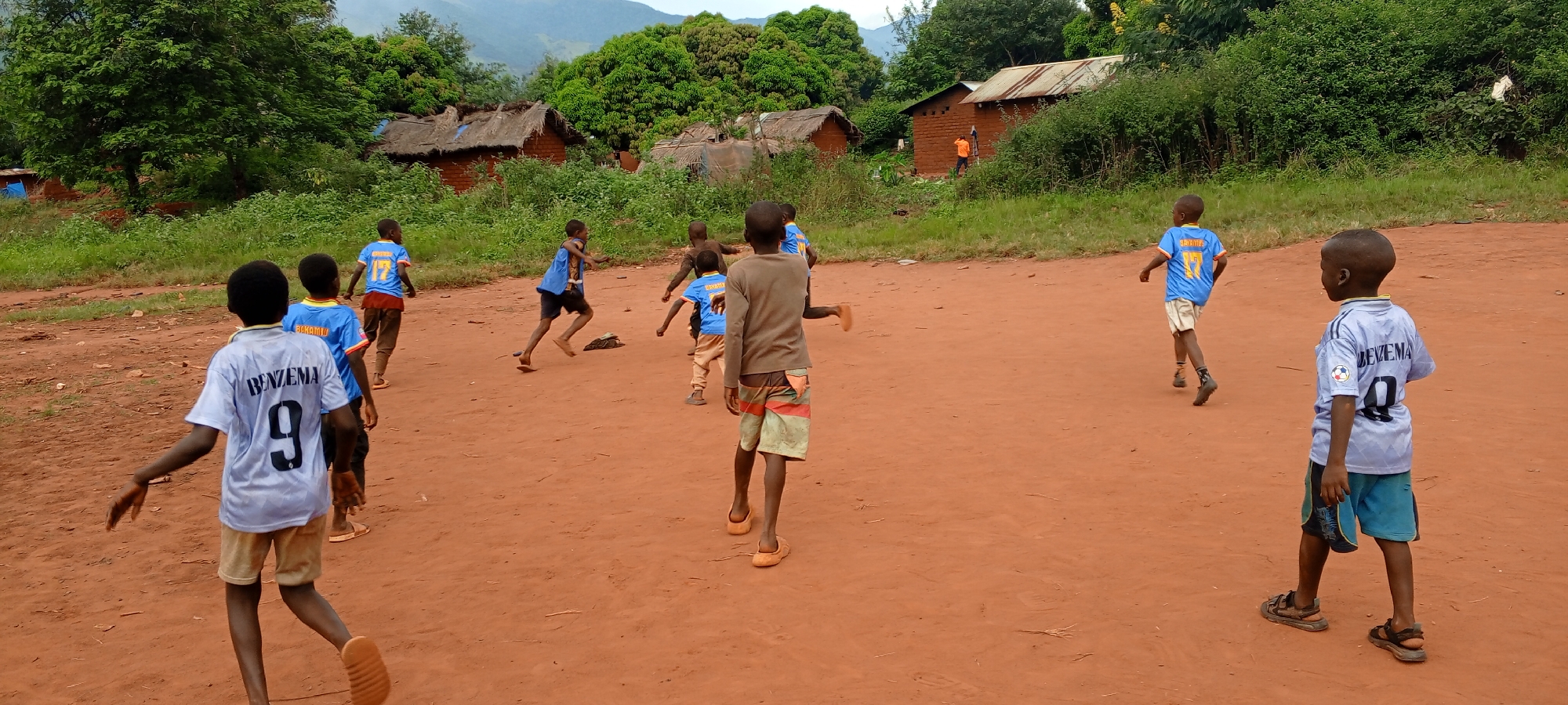 This dynamic image captures a group of young boys playing football on a reddish-brown dirt pitch in Ngalula, Democratic Republic of the Congo. Approximately eight to ten boys, mostly seen from behind, are scattered across the field, their postures indicating active movement in a game.
Many of the boys are wearing football jerseys. One boy in the immediate foreground wears a white jersey with "BENZEMA 9" printed in black on the back. Another boy further back wears a light blue jersey also with "BENZEMA 9." Other blue jerseys are visible, featuring names like "BAKAMBU 17" and "LUKAKU 17." Some boys are barefoot, while others wear sandals or flip-flops. Their expressions and body language convey the typical energy and enthusiasm of children at play.
The scene is set against a backdrop of simple, rural structures. Several single-story houses or huts, constructed with mud bricks or similar materials, have either thatched or corrugated iron roofs. These dwellings are surrounded by dense, lush green vegetation, including numerous trees and bushes, indicating a fertile environment. In the far distance, rolling hills or mountains are visible under a bright, overcast sky. The overall lighting suggests it is daytime, likely late morning or afternoon. The lack of harsh shadows implies a cloudy day. The image vividly portrays a moment of unadulterated childhood joy and activity within a humble, verdant community setting.