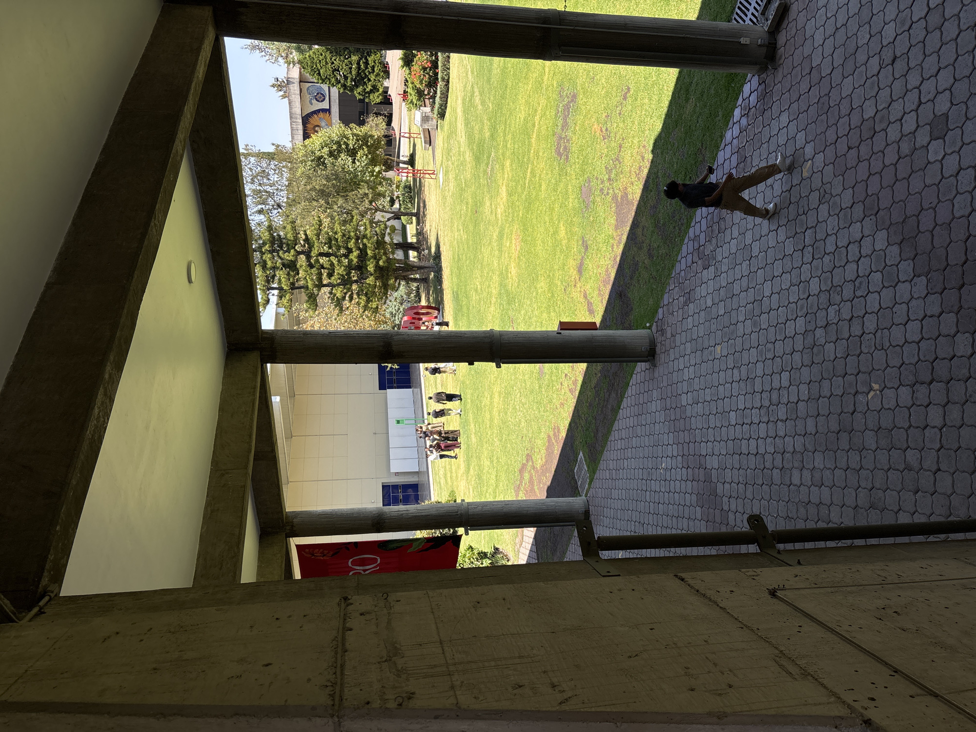 The image, taken in San Bernardino Tlaxcalancingo, Mexico, shows a view through a concrete structure, likely a building corridor or arcade. The scene is oriented at an angle. A grassy courtyard fills the center of the frame. In the distance, a building with a light-colored facade and blue window accents can be seen.
On the walkway to the right of the grass is a person wearing dark clothing and light-colored pants. A small group of people are gathered in the grass in the center of the courtyard. There are trees and other landscaping present in the courtyard.
The concrete structure frames the courtyard view, with concrete beams and pillars in the foreground. A banner is visible hanging from one of the pillars. The pavement is made of dark patterned tiles.