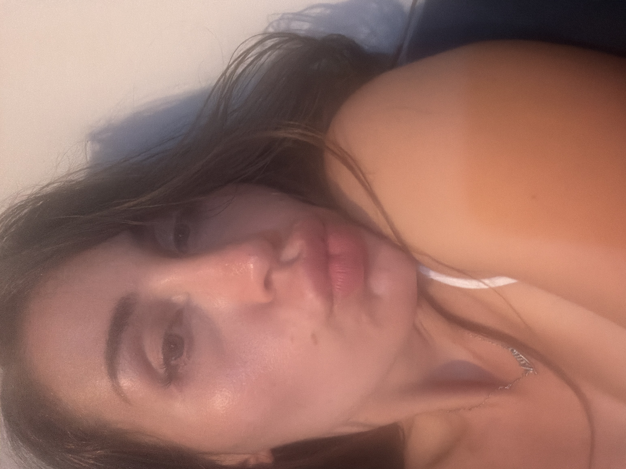 This is a close-up selfie of a woman with long, dark hair, taken indoors. She is leaning to her right, with her left shoulder visible. She is wearing a white top and a silver necklace with a nameplate. Her face is turned towards the camera, and her lips are pursed. The lighting is warm, casting a soft glow on her skin. The background is a plain, light-colored wall, with some shadows cast by what appears to be blinds or a similar object. There is no indication of the location being Tersefanou, Cyprus, or any other specific city.