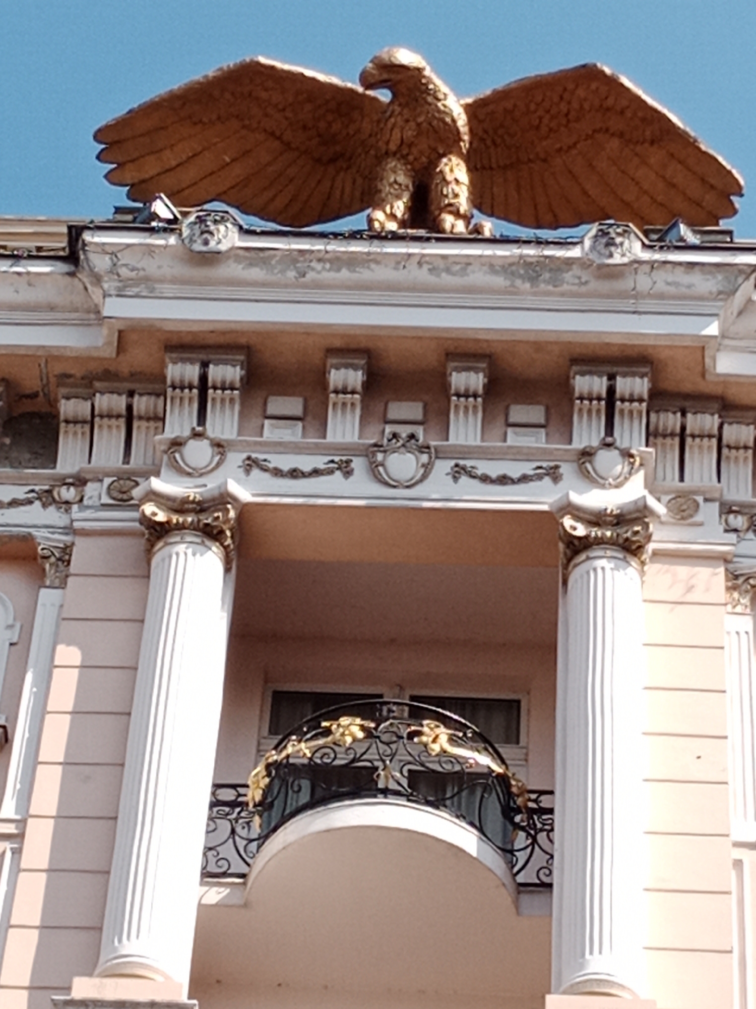 The image shows a close-up, low-angle shot of the upper facade of a building, possibly in Bydgoszcz, Poland. A large, golden eagle sculpture is perched on the rooftop, with its wings spread. Below the eagle, the building features ornate architectural details, including pilasters, decorative moldings, and columns. A curved balcony with black wrought-iron railings and gold embellishments is visible on a lower level. The building's exterior is painted a light pink or peach color, with white accents. The sky above is clear and blue. There are no people or animals visible in the image, and no discernible actions are taking place. The scene appears to be an exterior architectural detail of a historical building.