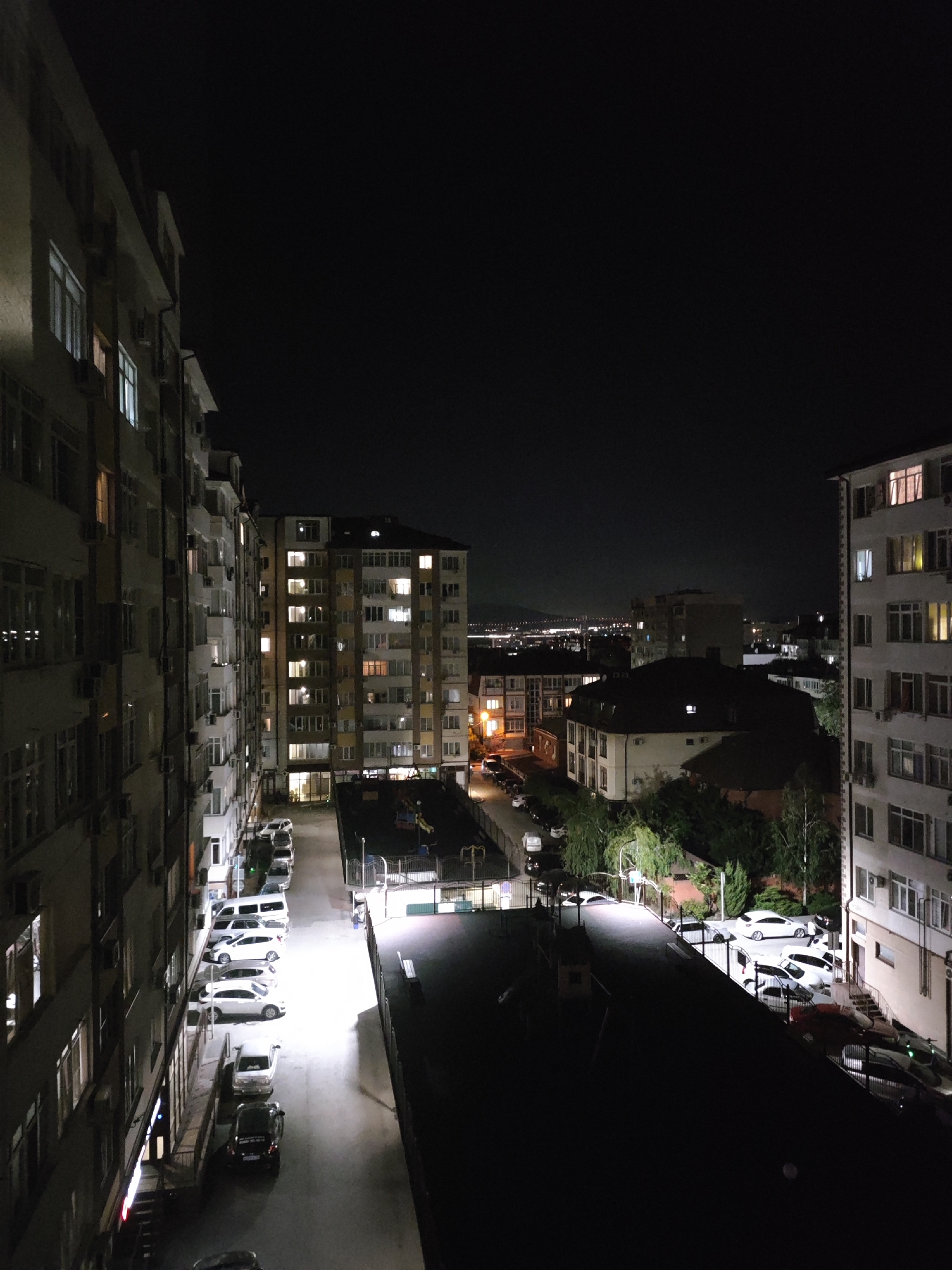 This is a nighttime aerial photograph of an urban residential area. In the foreground, a multi-story apartment building is visible on the left, with many illuminated windows. To the right, across a street, another apartment building is illuminated. Further in the background, more buildings and streetlights are visible, indicating a city environment. The sky is dark, and the scene is illuminated by artificial light sources. There are several cars parked along the street between the buildings. The location is identified as Gelendzhik, Russia. There are no people visible in the image.