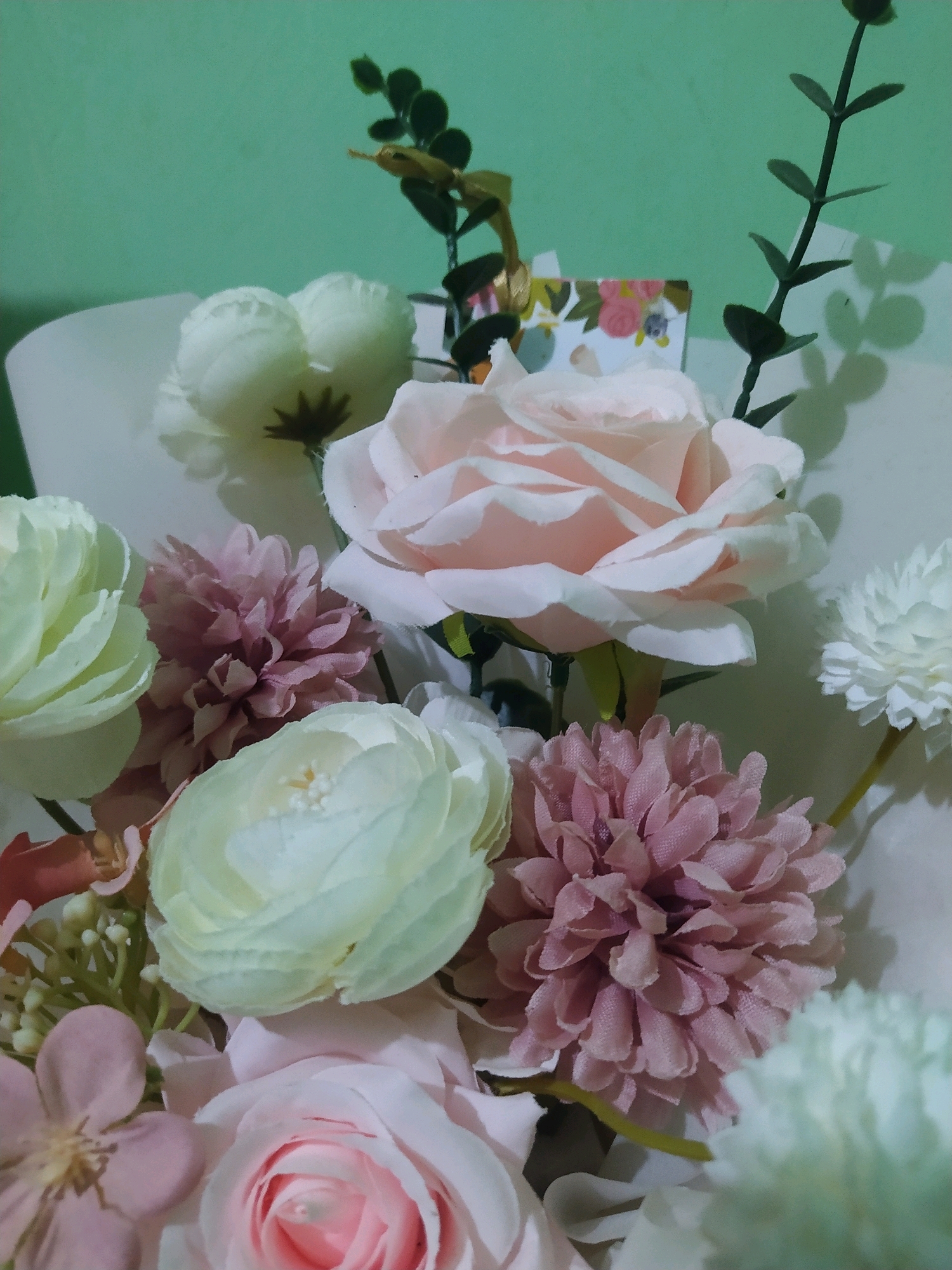 An arrangement of artificial flowers is presented against a pastel green wall. The bouquet includes several roses in pale pink and cream, along with pink and white pom-pom style flowers. Green artificial foliage and a small floral greeting card are also incorporated into the arrangement. The lighting casts soft shadows of the foliage onto the wall. The scene appears to be indoors, with the arrangement likely placed on a surface out of frame. There is no indication of the city of Sroyo, Indonesia in the image itself.