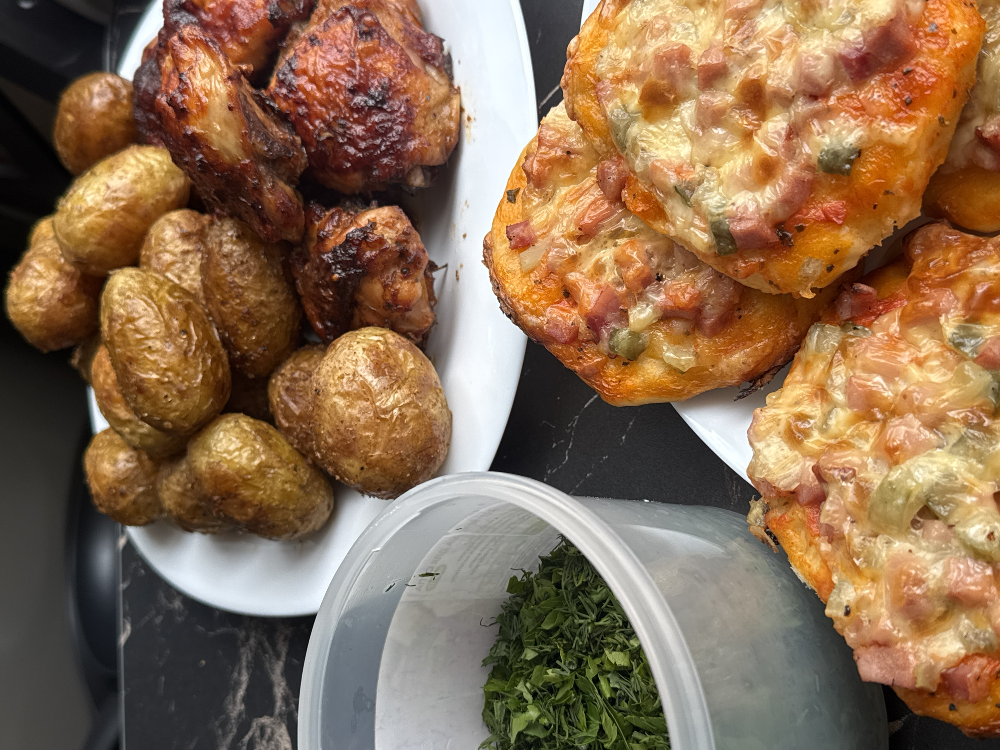 The image depicts a meal, likely homemade, consisting of roasted chicken pieces, small roasted potatoes, and mini pizzas with ham and cheese topping. A plastic container with chopped herbs, possibly parsley or dill, is also present. The scene appears to be set on a dark, possibly marble-patterned surface, suggesting a kitchen or dining area. The location is Chaikovsky, Russia, but there are no contextual clues in the image itself to specifically identify this city. Therefore, it is simply a close-up shot of food. There are no people visible, and the focus is solely on the presentation of the food.