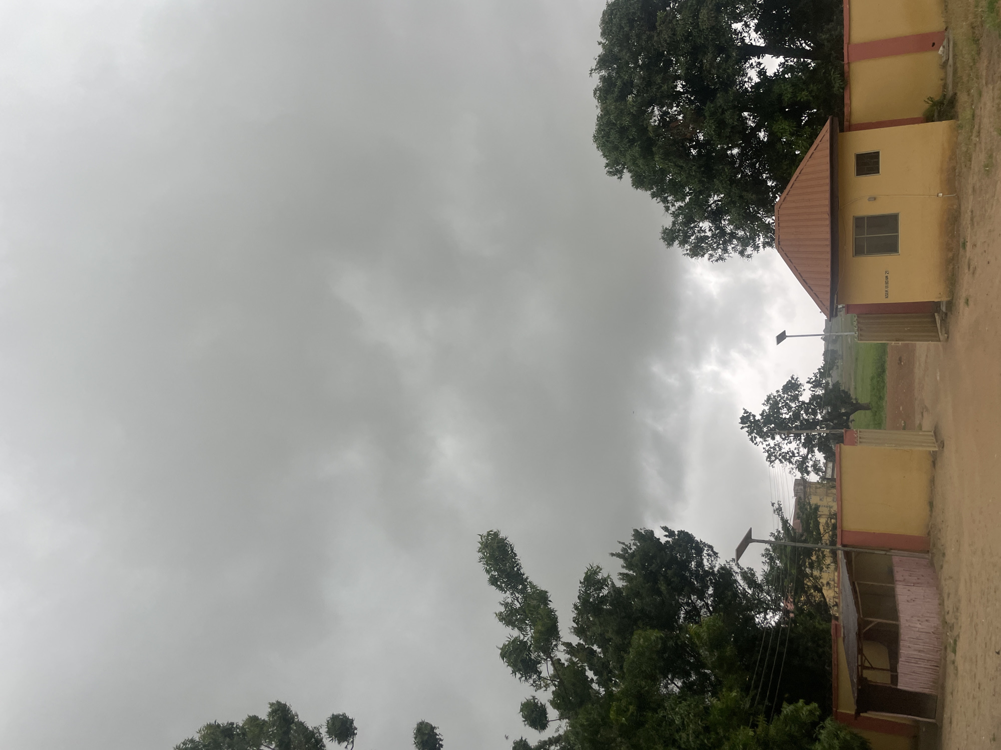 The image depicts a slightly overcast day in Keffi, Nigeria, with a focus on a gated entrance to what appears to be a residential or administrative compound. The sky is filled with heavy, gray clouds, suggesting an impending rain or a generally gloomy atmosphere.
In the foreground, a sandy path leads to the entrance. To the left, a small, open-sided structure with a thatched roof is visible, constructed with wooden posts and pinkish bamboo or reed walls. Adjacent to this is a section of a yellow wall with a red trim, forming part of the compound's perimeter. A tall, slender lamp post with a solar panel stands near this structure.
To the right, a more substantial building with a brown, corrugated metal roof is situated. This building has two windows and a sign with text that is partially legible, reading "NSUK GURUN 21." This building also features yellow walls with red trim, consistent with the other visible boundary walls. A metal gate, partially ajar, provides access through the wall.
The background reveals lush green trees, some quite large and mature, framing the scene. Beyond the compound, a field of green grass stretches into the distance, with more trees dotting the landscape under the muted sky. The overall impression is one of a semi-tropical environment under a threatening sky. There are no people visible in the image.