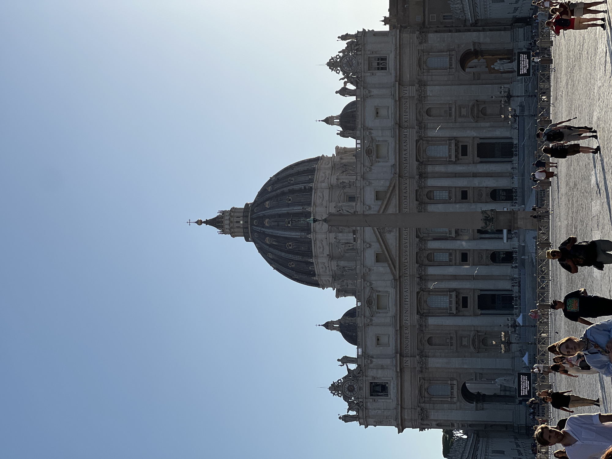 This full vertical shot captures St. Peter's Basilica and a portion of St. Peter's Square in Vatican City, Rome, Italy, under a clear, light blue sky. The grand facade of the basilica dominates the upper background, featuring multiple levels of classical architecture, columns, and a large central dome topped with a cross. Statues line the roofline, and inscribed Latin text, "IN HONOREM PRINCIPIS APOST PAVLV SV BVRGH," is visible above the main entrance area.
In the mid-ground, an ancient Egyptian obelisk stands vertically, partially obscuring the lower facade. The square's paved surface is visible, covered with numerous individuals walking in various directions. Most people are dressed in casual summer attire, and many cast long shadows to their left, indicating the sun is positioned to the right of the frame, likely late in the day. Metal crowd control barriers delineate sections of the square, and a large dark sign with white text is visible near the base of the basilica on the left side.