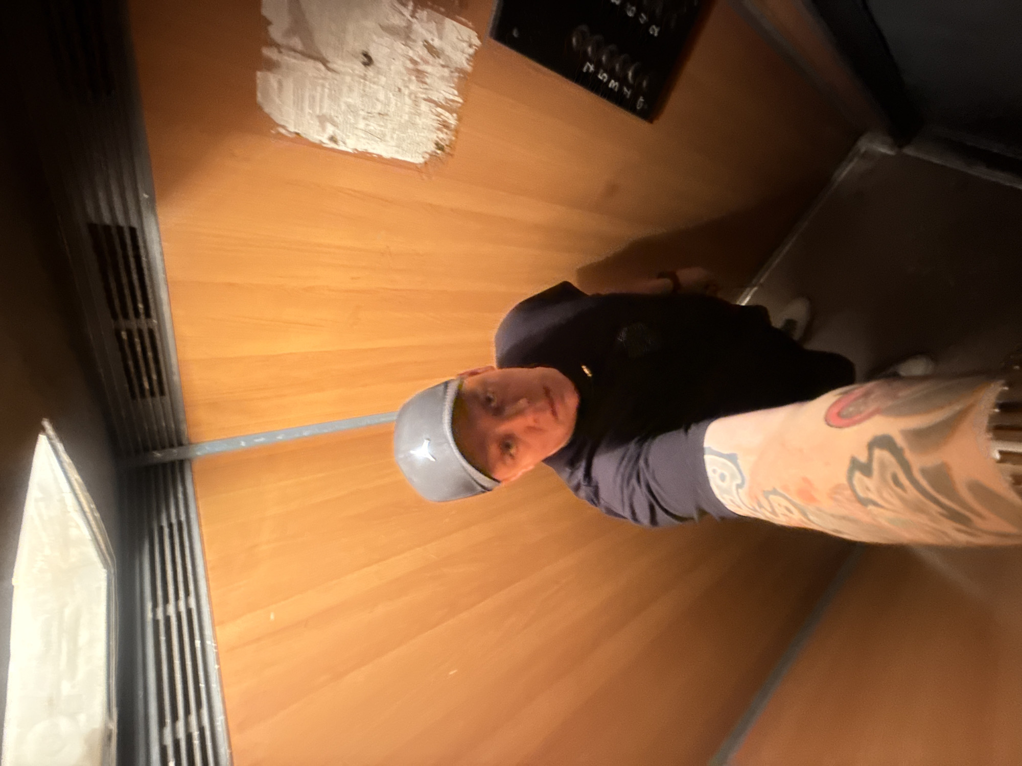 A person wearing a black t-shirt and a grey baseball cap is standing in an elevator with wood-paneled walls. The person is holding a phone to take a selfie, and their left arm, which is heavily tattooed, is visible in the foreground. An elevator control panel with buttons is to the right of the person. The elevator appears to be old and somewhat worn, with a patch of white material on the wall. The overall scene suggests a candid or informal moment captured within the elevator. The location is not discernible from the image itself.