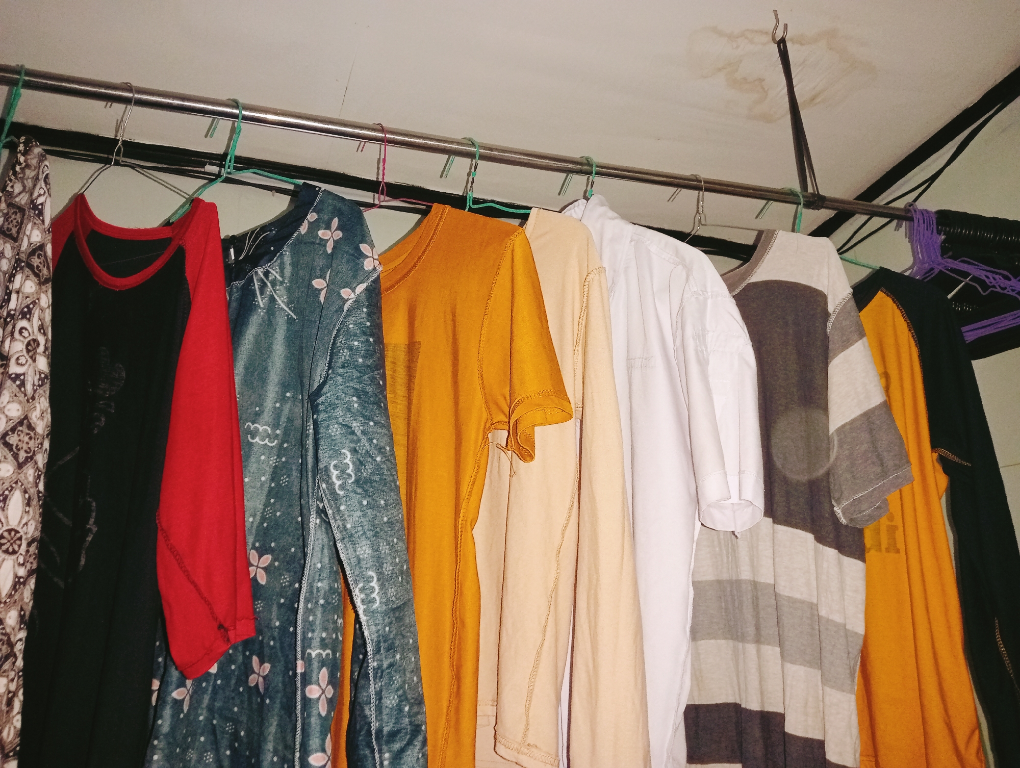 The image depicts an indoor clothing rack displaying several garments hanging from green and pink plastic hangers. The garments include a patterned fabric, a black shirt with red sleeves, a blue-green patterned dress, an orange t-shirt, a tan t-shirt, a white t-shirt, a gray and white striped t-shirt, and a yellow t-shirt. The clothing rack is suspended from the ceiling, which appears to be stained in several areas. The scene is indoors, suggesting it could be within a residential or commercial space in Tridayasakti, Indonesia, possibly a laundry area or a storage space for clothing. There are no people visible in the image. The lighting is somewhat dim, casting shadows on the clothes and the ceiling.