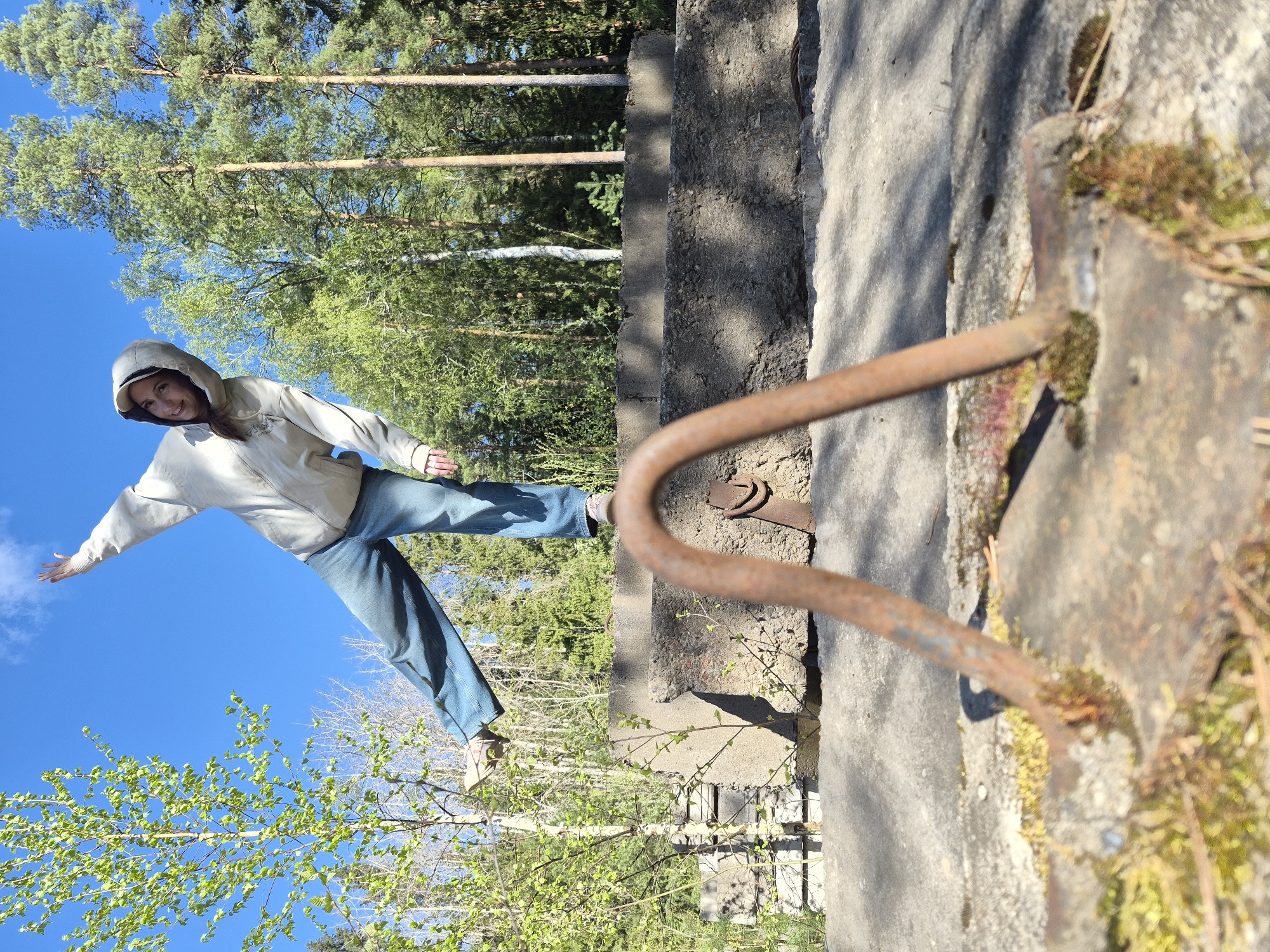 A young woman stands atop a weathered concrete structure in a wooded area, possibly in Chaikovsky, Russia. She is balanced with one leg extended outward and arms outstretched. She wears a light-colored hoodie, blue jeans, and sneakers. The sky is clear blue, punctuated by scattered clouds and surrounding trees. A rusty, curved metal railing is visible on the right side of the frame, attached to the concrete structure. Moss grows on the structure, indicating age and exposure to the elements. Vegetation grows below, obscuring what the concrete structure sits on.