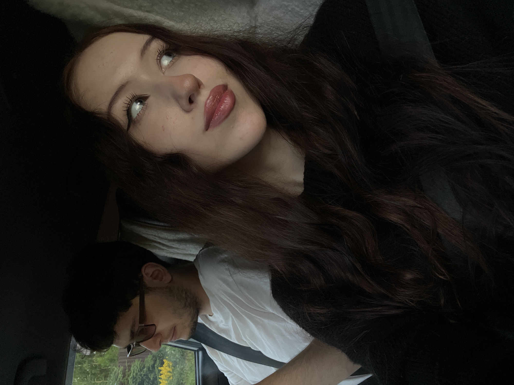 In a car, a young woman with long, dark, wavy hair and dark eye makeup is visible in the foreground, looking to the right. She is wearing a black top and a seatbelt. Behind her, to the left, a young man with dark hair and glasses is wearing a white t-shirt and a seatbelt. He is looking to the left. The interior of the car is dimly lit, with some natural light coming through the windows, revealing greenery and a wooden fence outside. The image is a selfie taken by the woman. The location is likely Votkinsk, Russia, based on the user's prompt.